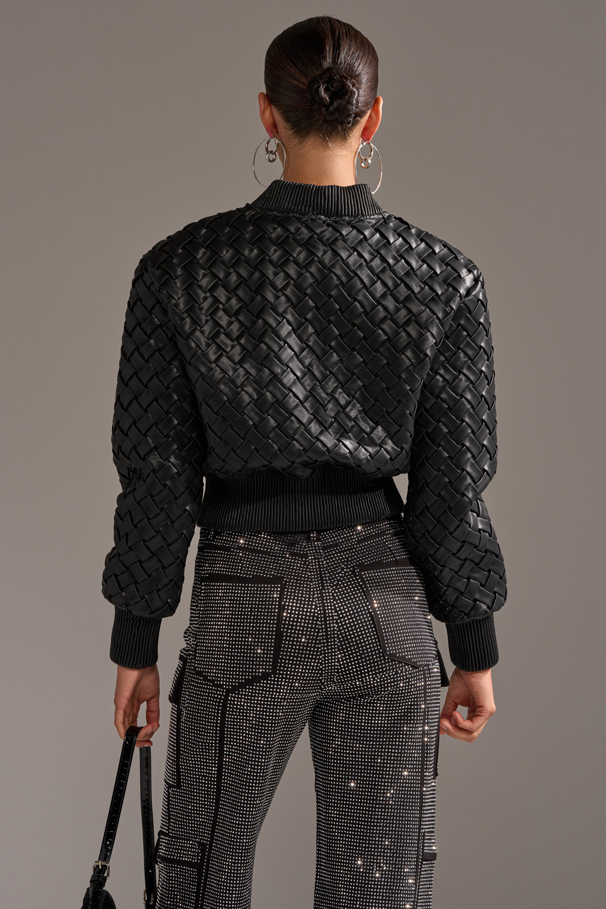 A person with a bun, wearing the WICKER PARK WOVEN BOMBER and embellished pants, is shown from behind holding a black handbag against a plain, neutral background.