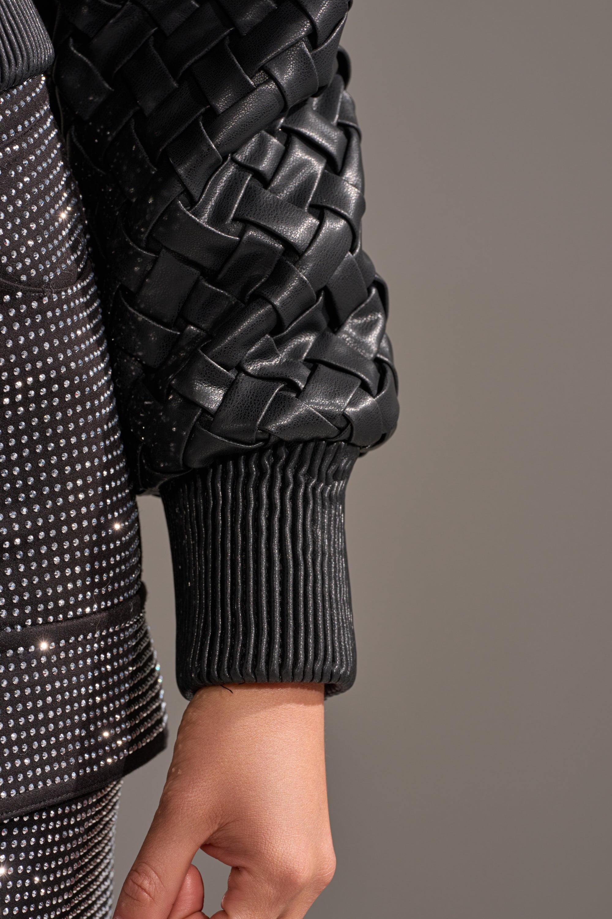 A close-up of an arm in the WICKER PARK WOVEN BOMBER, a textured black jacket with ribbed cuffs, appears beside a black studded garment on a neutral gray background.