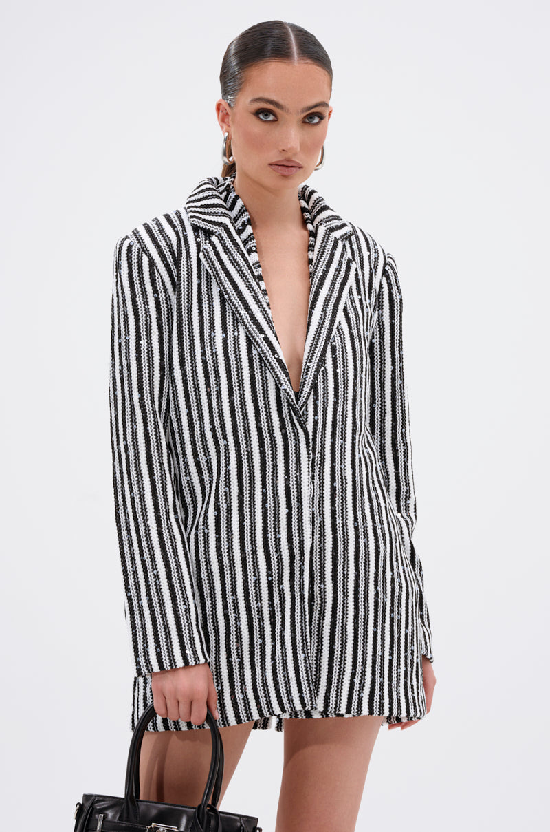 A woman with slicked-back hair wears the NORTH AVE CROCHET BLAZER, a black and white vertically striped knit blazer dress. She holds a black handbag, has bold dark eyeliner makeup, and poses against a plain white background.