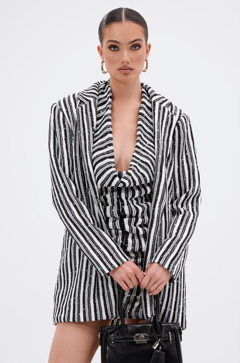 A woman with slicked-back hair wears the NORTH AVE CROCHET BLAZER dress with a deep neckline, holding a black handbag. She accessorizes with hoop earrings and poses confidently against a plain white background.