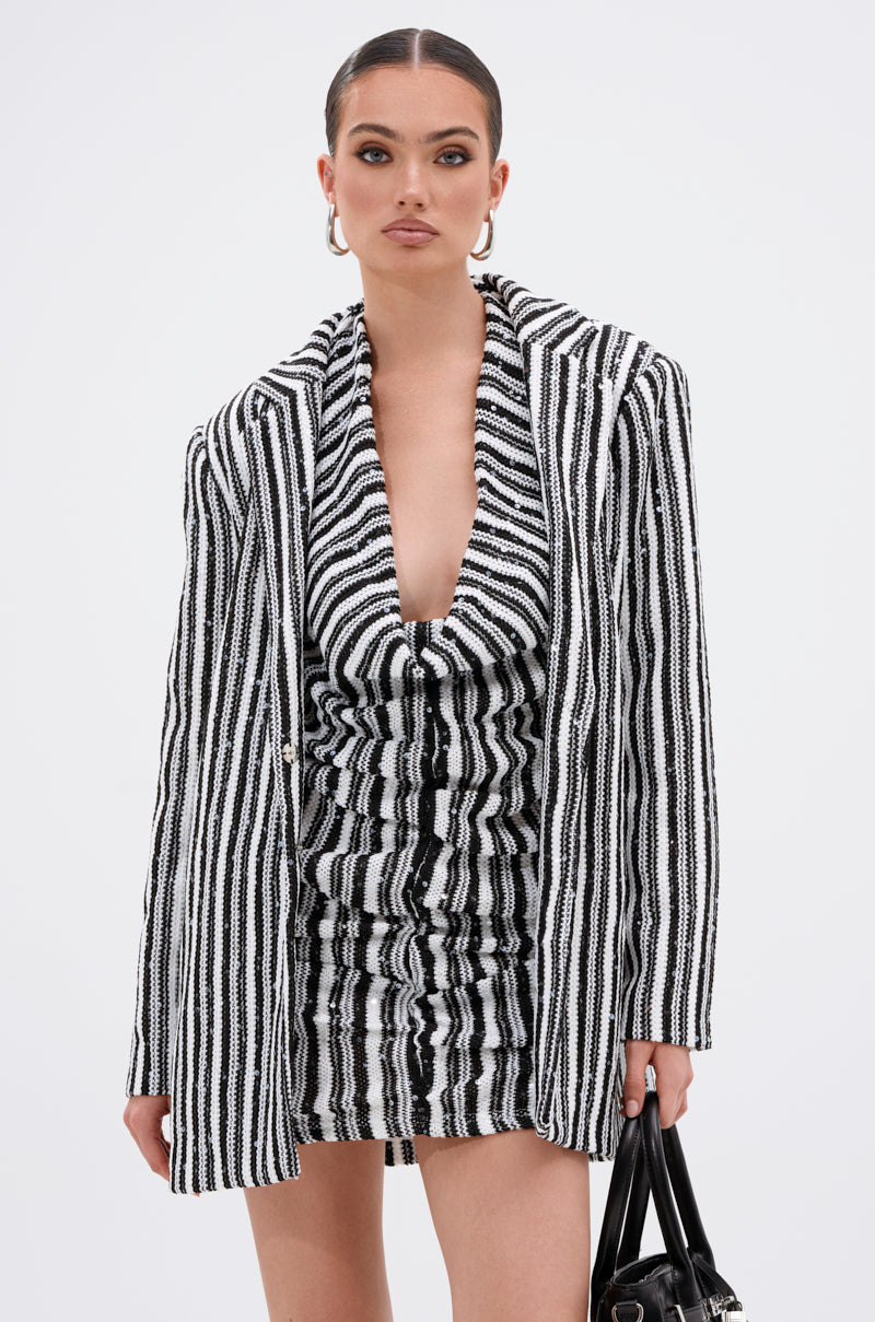 A woman stands against a plain white background holding a black handbag, wearing the NORTH AVE CROCHET BLAZER over a black and white striped ruched knit dress.
