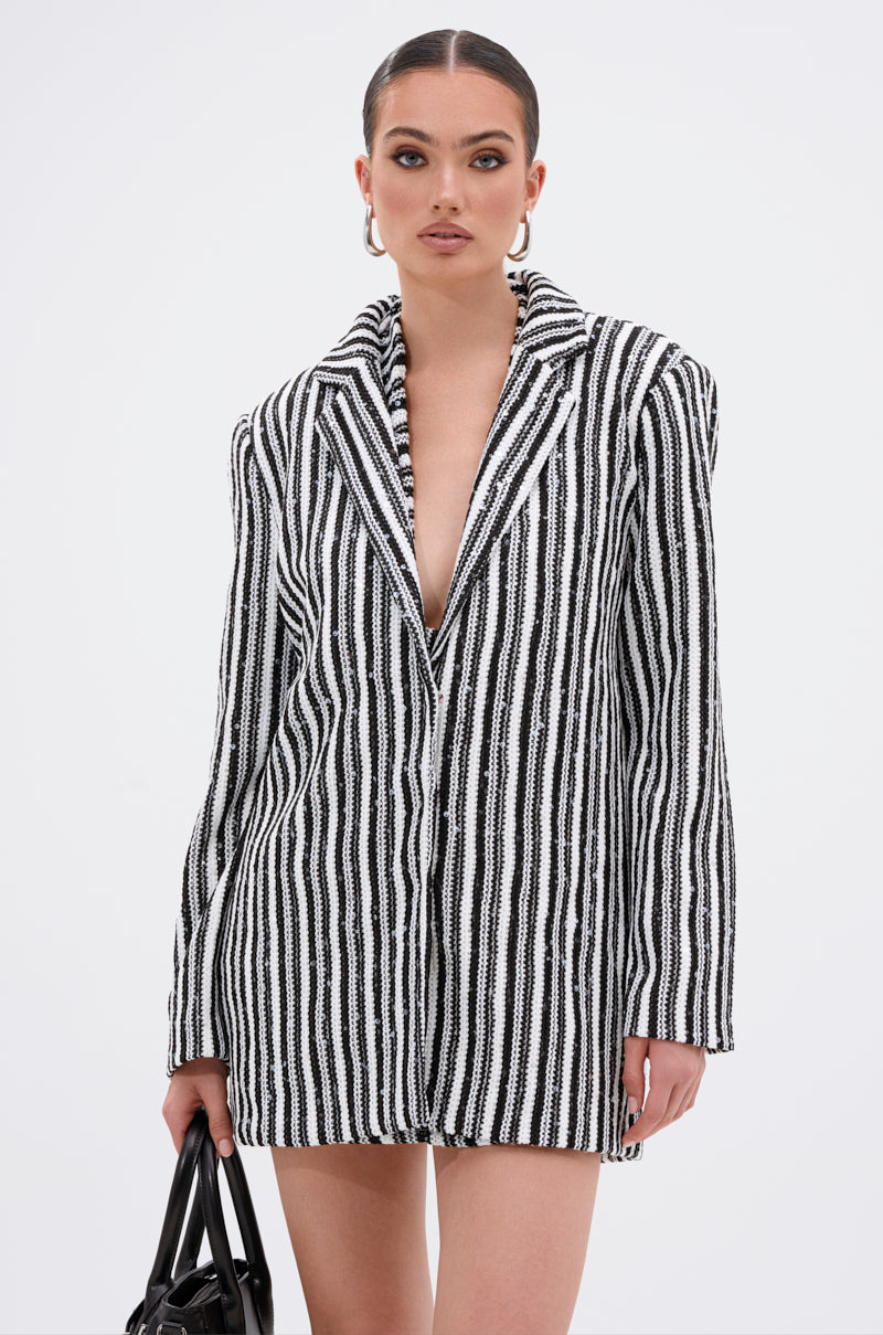 A woman poses against a white background wearing the NORTH AVE CROCHET BLAZER, a black and white vertically striped knit oversized blazer styled without a shirt. She accessorizes with hoop earrings, slicked-back hair, and carries a black handbag.