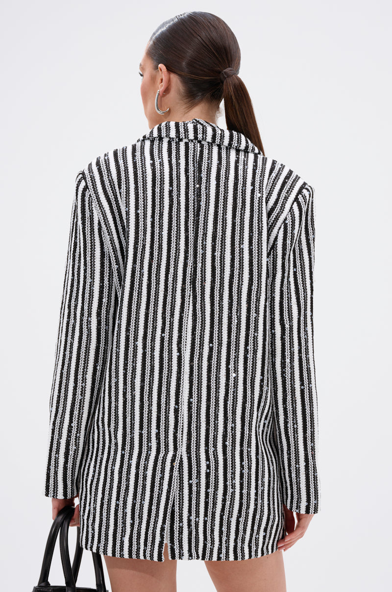 A woman with straight brown hair in a low ponytail wears the NORTH AVE CROCHET BLAZER in black and white and holds a black handbag, shown from the back against a plain white background.