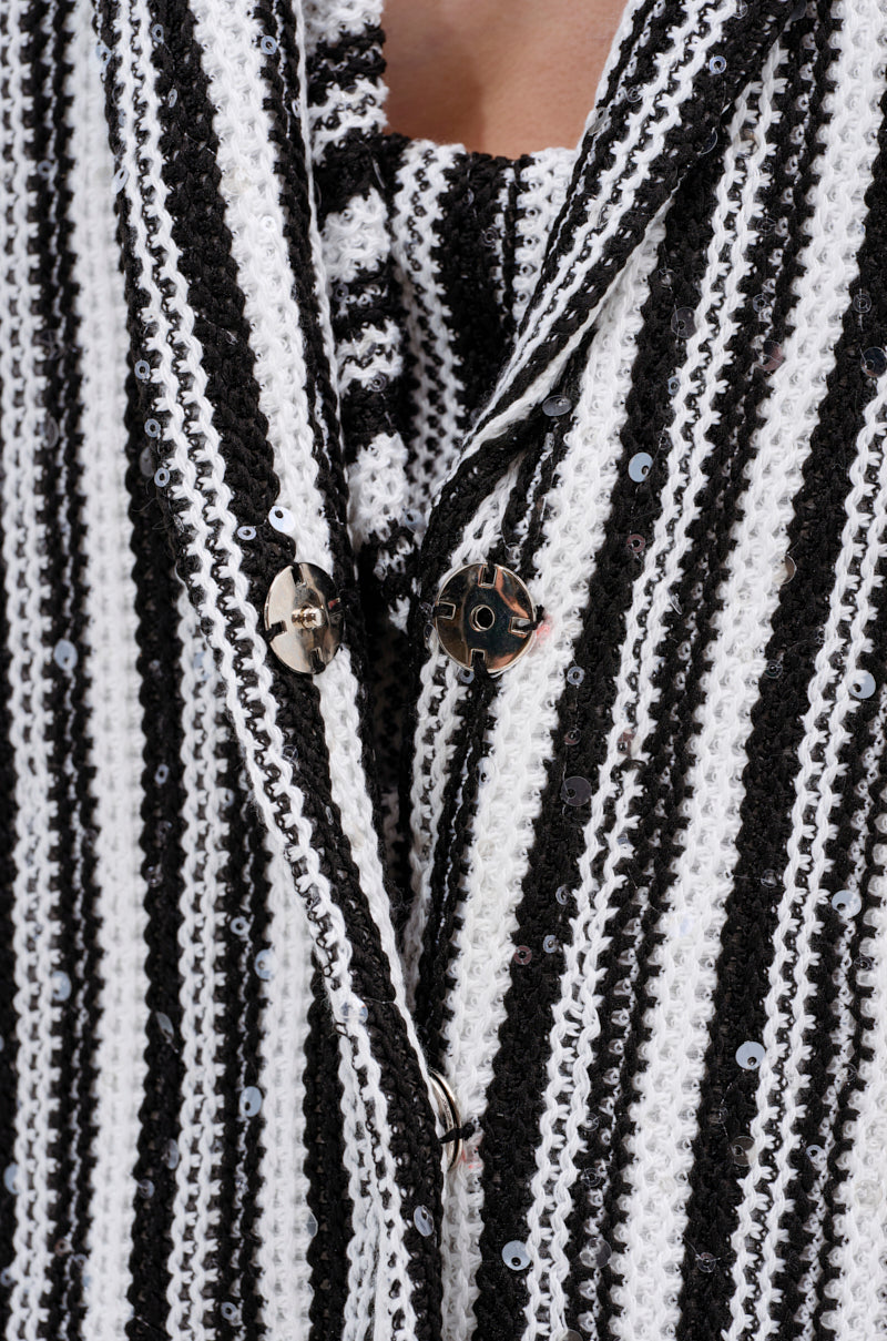Close-up of the NORTH AVE CROCHET BLAZER, a black and white striped knit jacket with sequin details and metallic round buttons, worn over a matching top. The fabric has a woven, textured pattern.