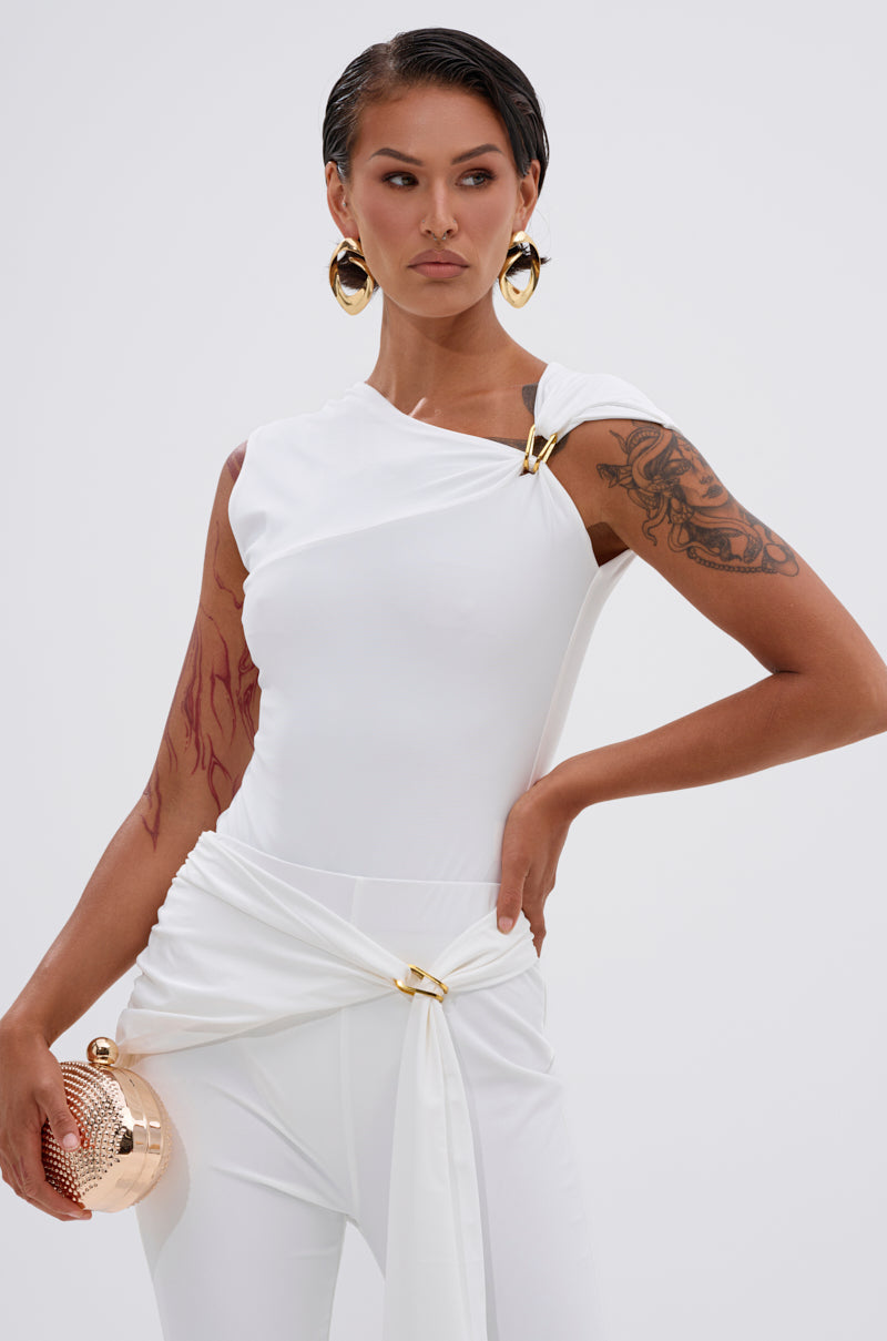 A woman with slicked-back hair, large gold hoop earrings, and tattoos poses in the body-contouring IZZY SEAMLESS BODYSUIT, holding a round gold clutch against a plain light background. 1