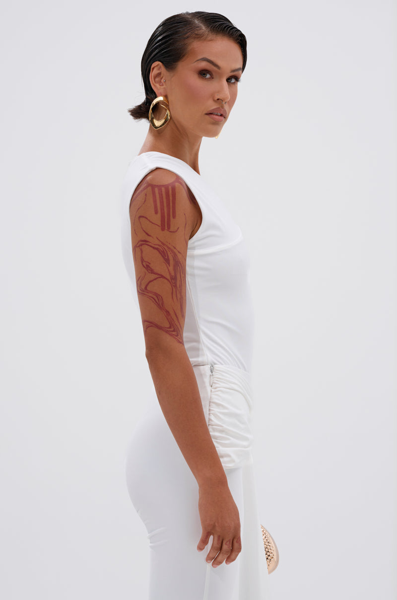 A woman with slicked-back hair, gold hoop earrings, and a red upper-arm tattoo models the fitted IZZY SEAMLESS BODYSUIT, highlighting its contouring fabric as she stands in profile before a plain light background. 3