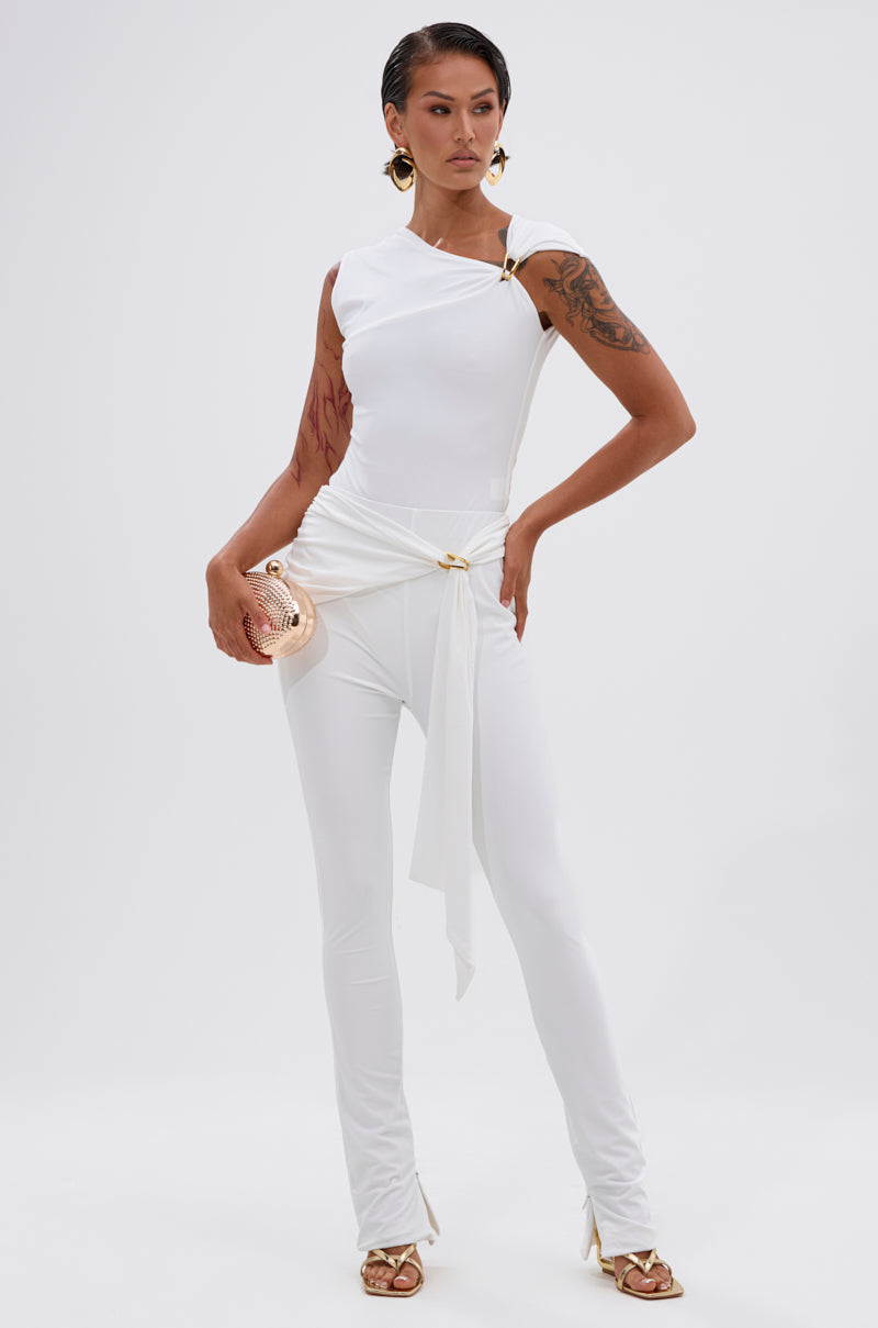A woman poses confidently in the IZZY SEAMLESS BODYSUIT, a stylish fitted white piece with gold accents and body-contouring fabric. She holds a metallic clutch, has slicked-back hair, visible tattoos, and wears strappy heels against a plain white background. 7