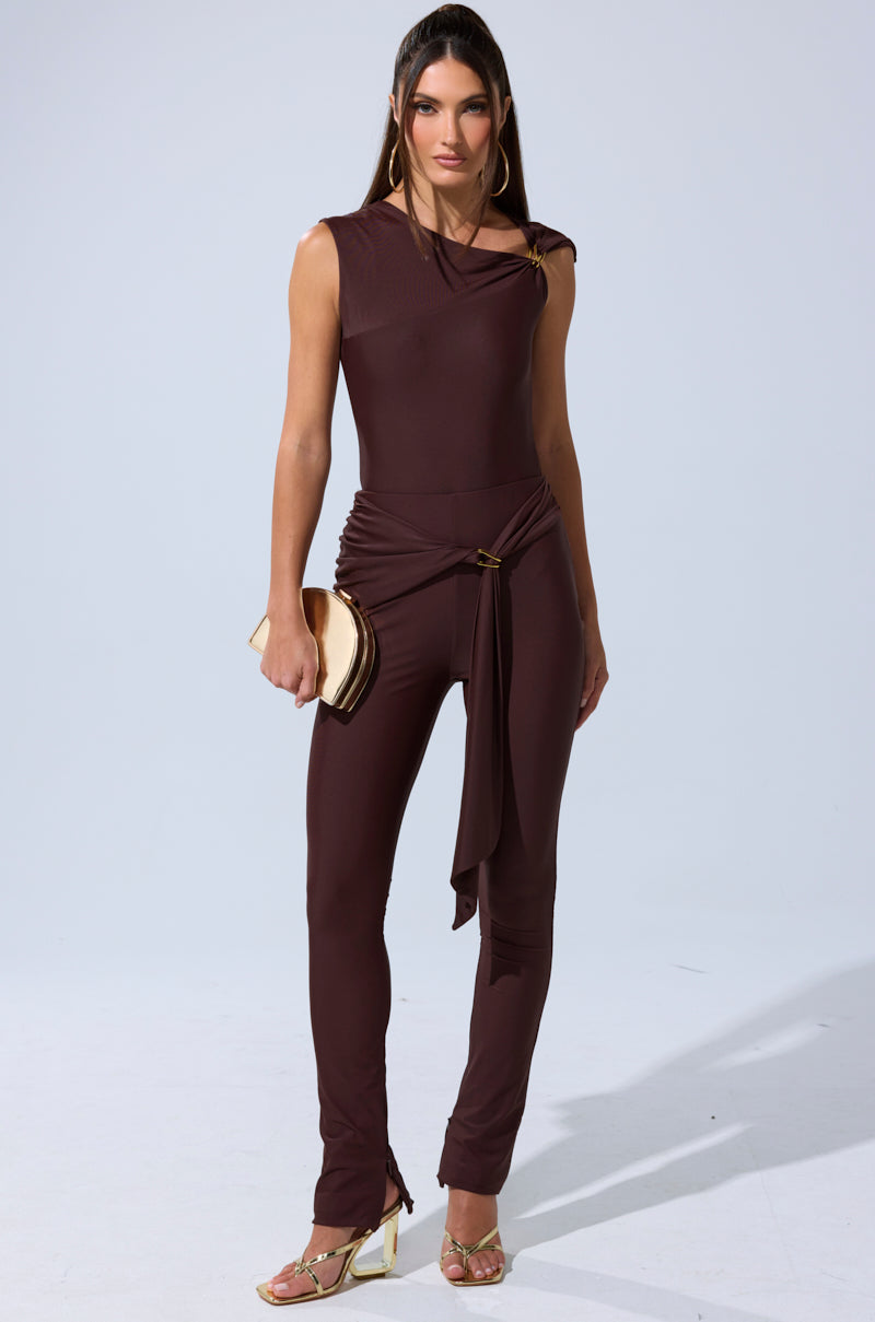A woman with long, straight hair and a neutral expression stands against a plain background, wearing the KIMMY SEAMLESS BODYSUIT in dark brown with an asymmetric neckline and draped waist tie, paired with gold heels and a beige clutch. 9