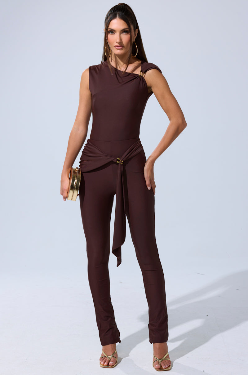 A woman faces forward wearing the KIMMY SEAMLESS BODYSUIT—a fitted, stretch-knit jumpsuit with an asymmetric neckline and belt detail. She accessorizes with a gold clutch, gold hoop earrings, and heeled sandals against a plain light background. 8