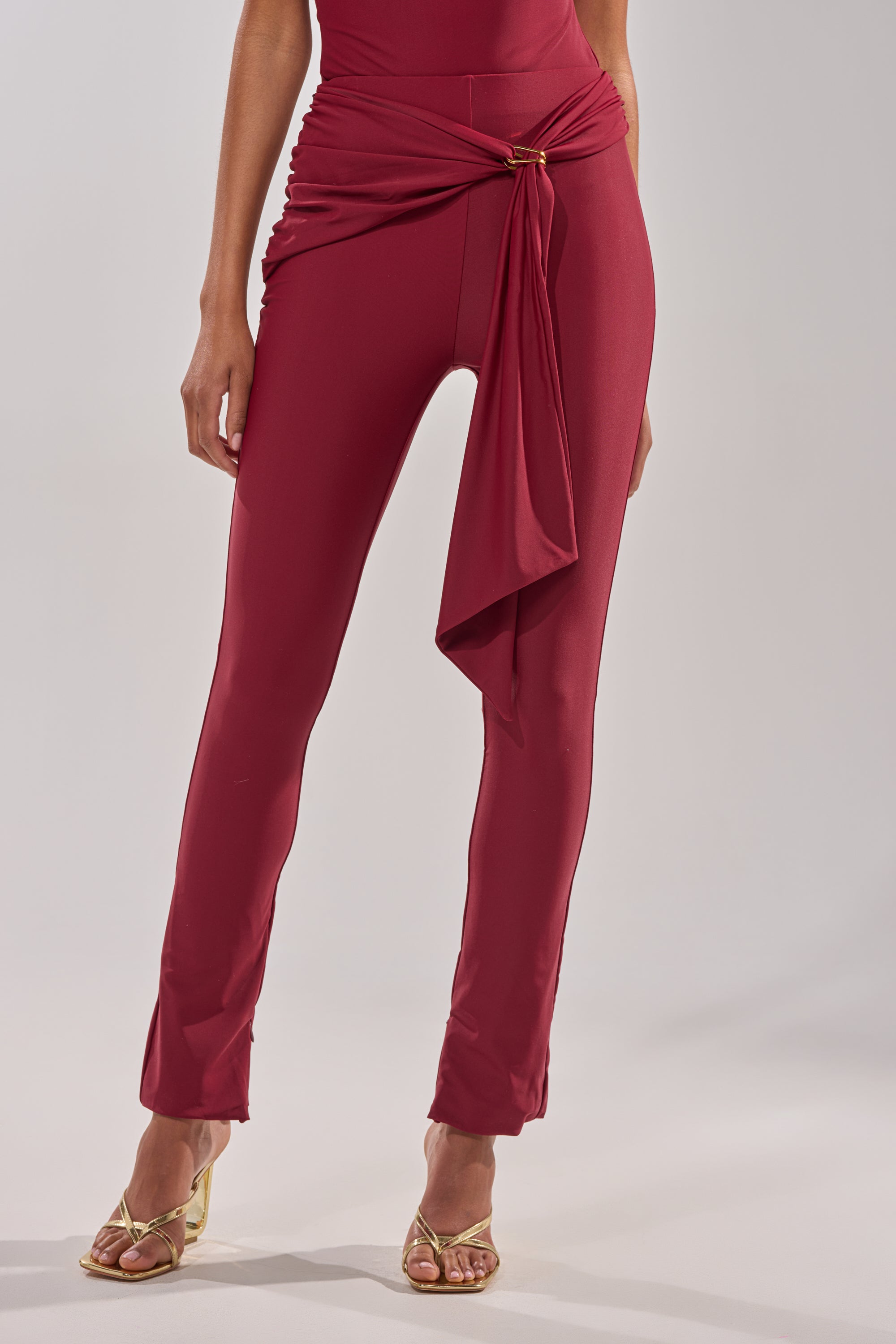 A person wears the IZZY SEAMLESS LEGGING IN BURGUNDY, a high-waisted, fitted burgundy pant, paired with gold open-toe heeled sandals. The upper body and face are not visible. 1