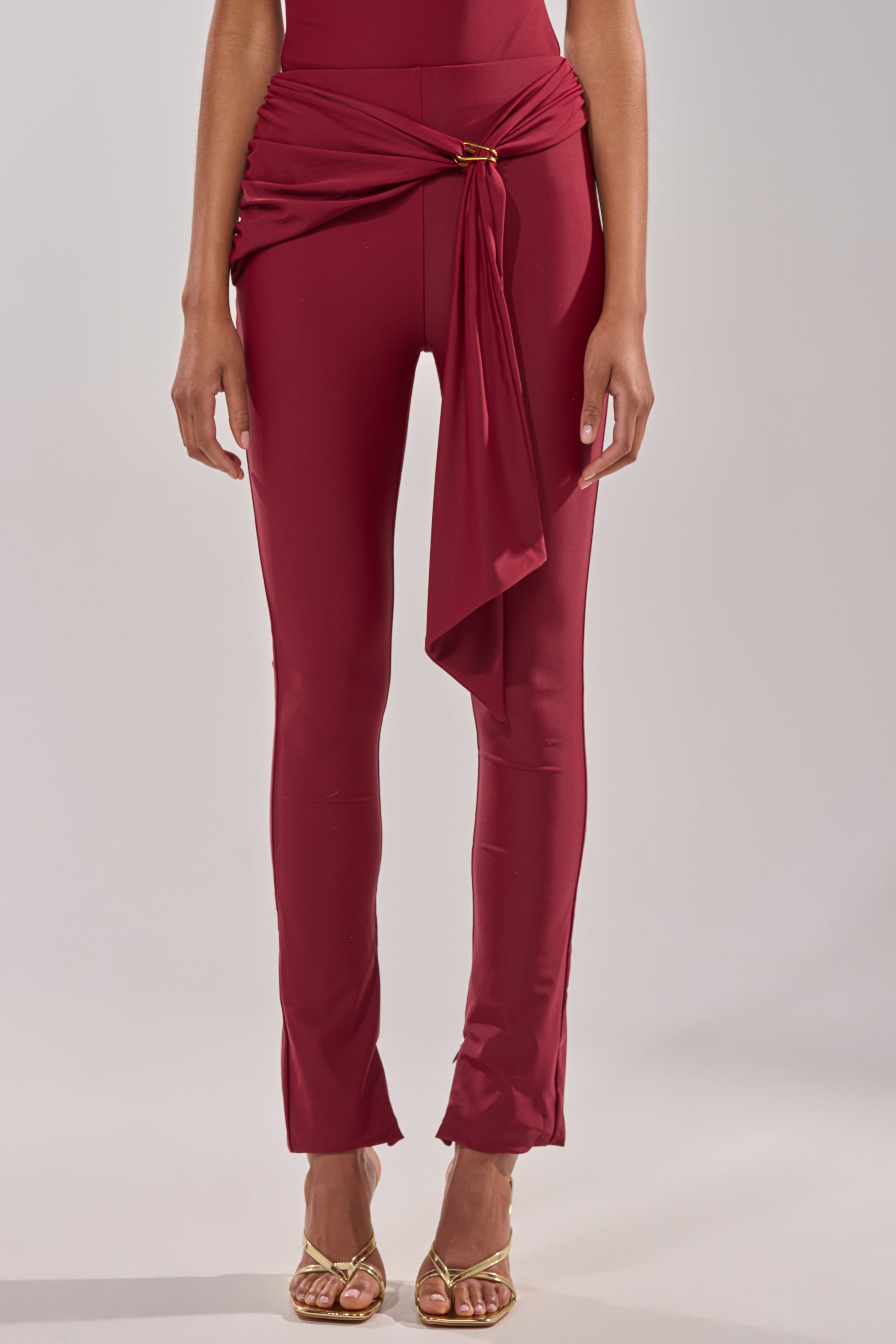 A person wears the IZZY SEAMLESS LEGGING IN BURGUNDY, styled with a draped sash at the waist and paired with gold strappy high-heeled sandals, set against a plain neutral background. 2