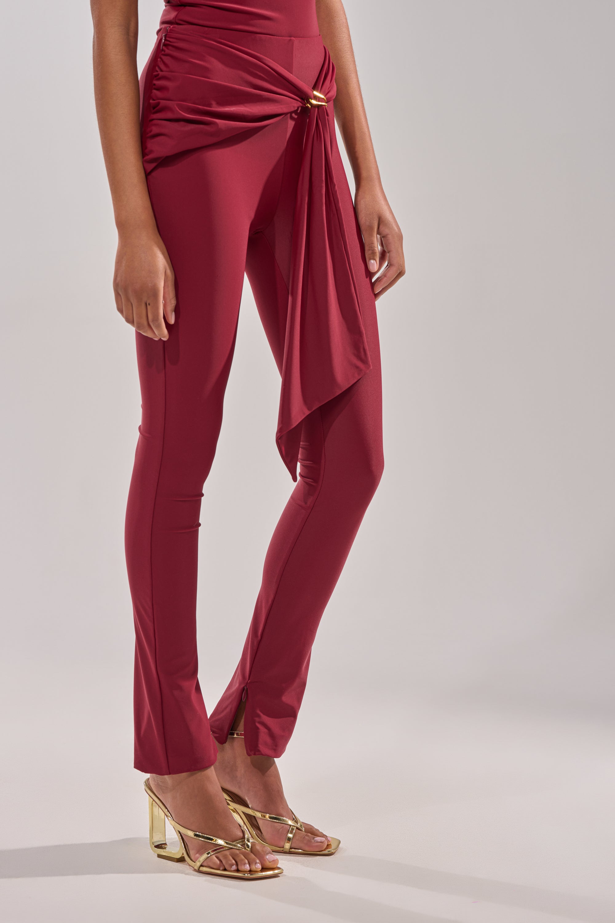 A person in the IZZY SEAMLESS LEGGING IN BURGUNDY and gold heeled sandals stands against a plain light background. 4
