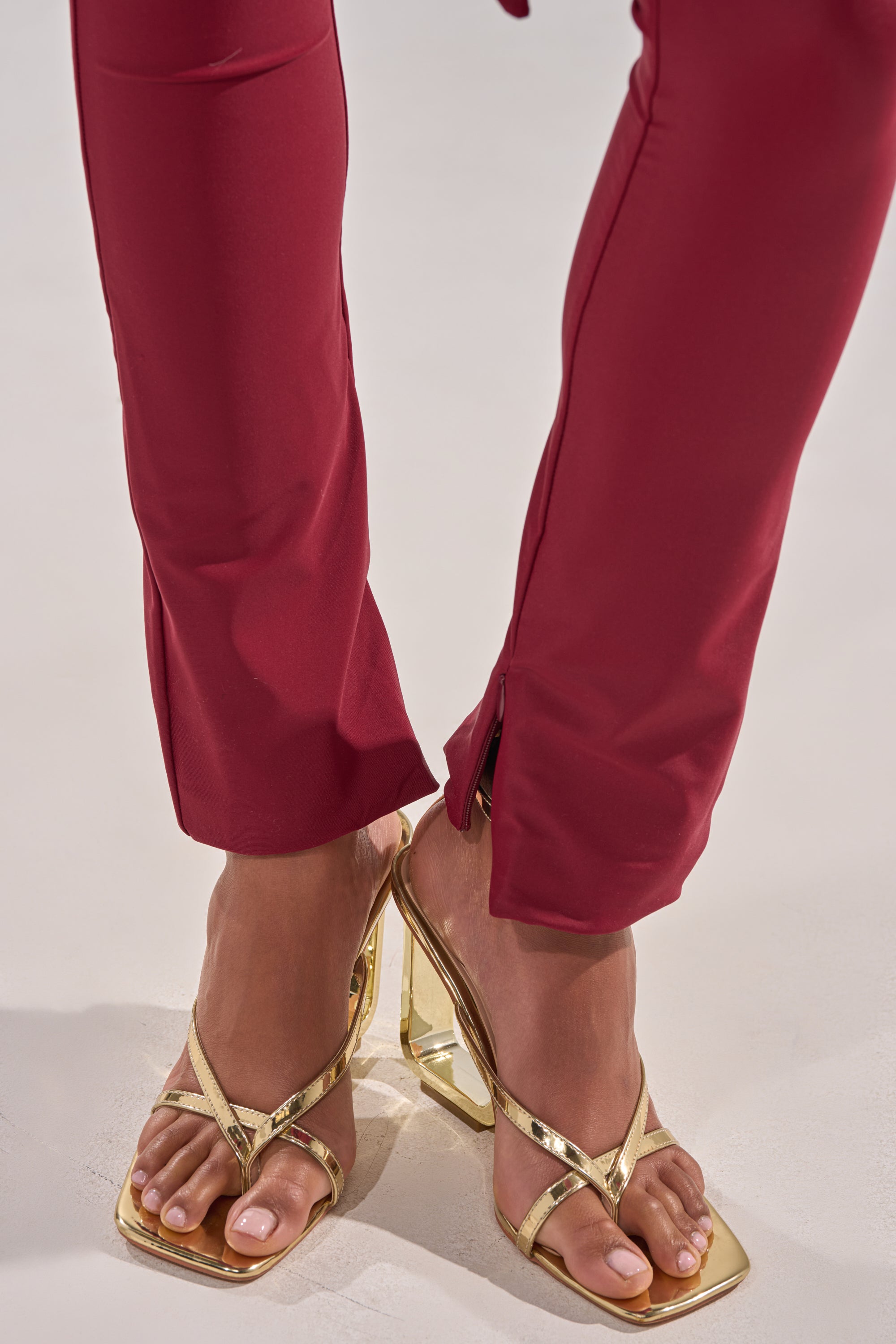 Close-up of a person wearing the IZZY SEAMLESS LEGGING IN BURGUNDY with front hem slits and gold high-heeled sandals, standing on a light-colored surface. 8