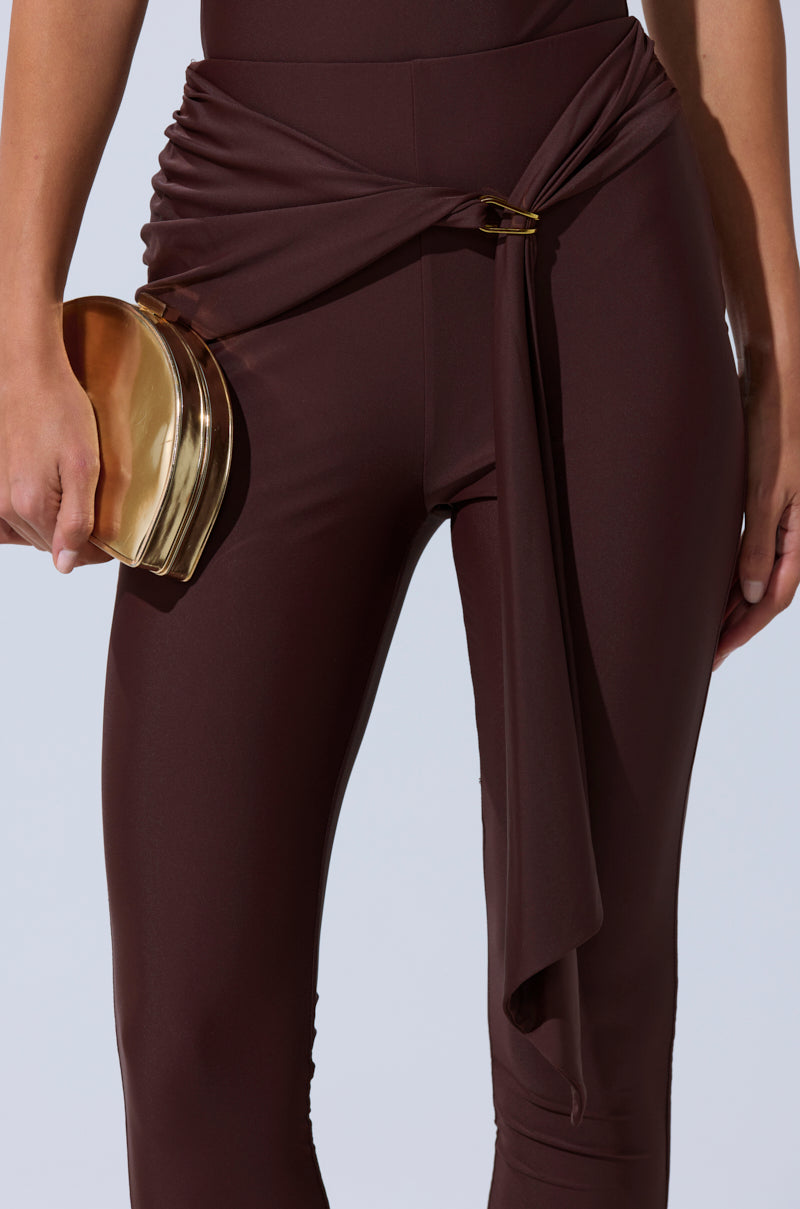 Wearing the KIMMY SEAMLESS LEGGING IN BROWN, featuring a high-waisted, body-contouring fit and draped belted detail, paired with a small gold metallic clutch in the left hand. 9
