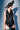 A person wearing the KRISTIN SATIN BLOUSE, black lace shorts, thigh-high black boots, and a wide-brimmed black hat with decorative trim, standing against a light blue background.