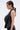 A woman with slicked-back hair is shown in profile wearing the KRISTIN SATIN BLOUSE, a black, sleeveless, asymmetrical top with a tie-back detail, paired with black lace bottoms against a plain background.