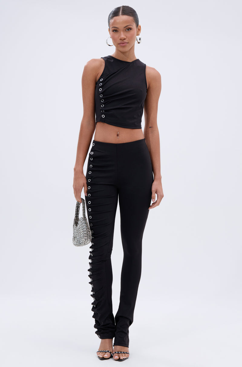 A woman stands against a plain background in a sleeveless black crop top with silver grommet accents and the GROMMET DETAIL RUCHED PANT—high-rise, curve-hugging skinny pants. She accessorizes with a silver bag and sandals. 1