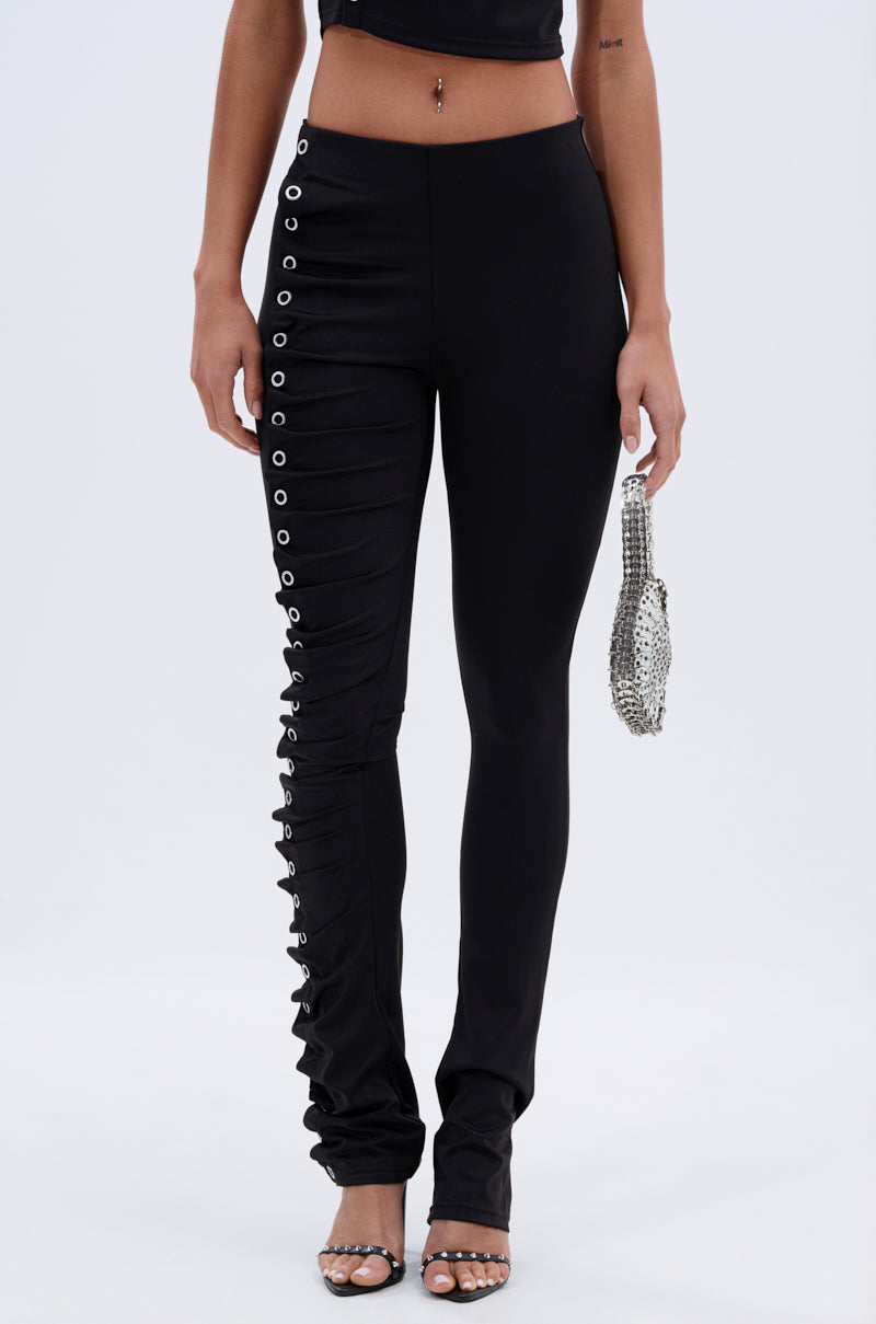 A woman wears GROMMET DETAIL RUCHED PANT with silver grommets on one leg, a black crop top, silver high-heeled sandals, and carries a small silver beaded bag. 0