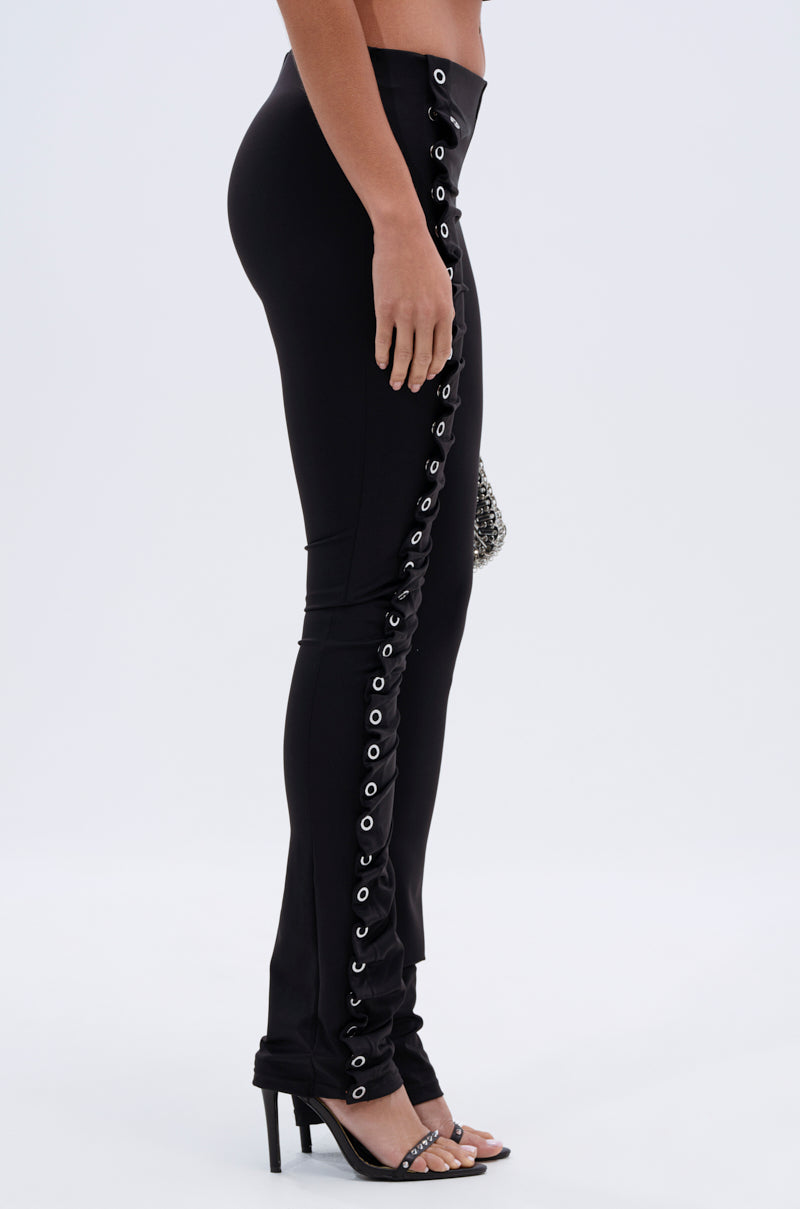 Someone models the GROMMET DETAIL RUCHED PANT, featuring high-rise skinny legs with silver grommet eyelets and side lacing, styled with black heeled sandals against a plain light background. 2