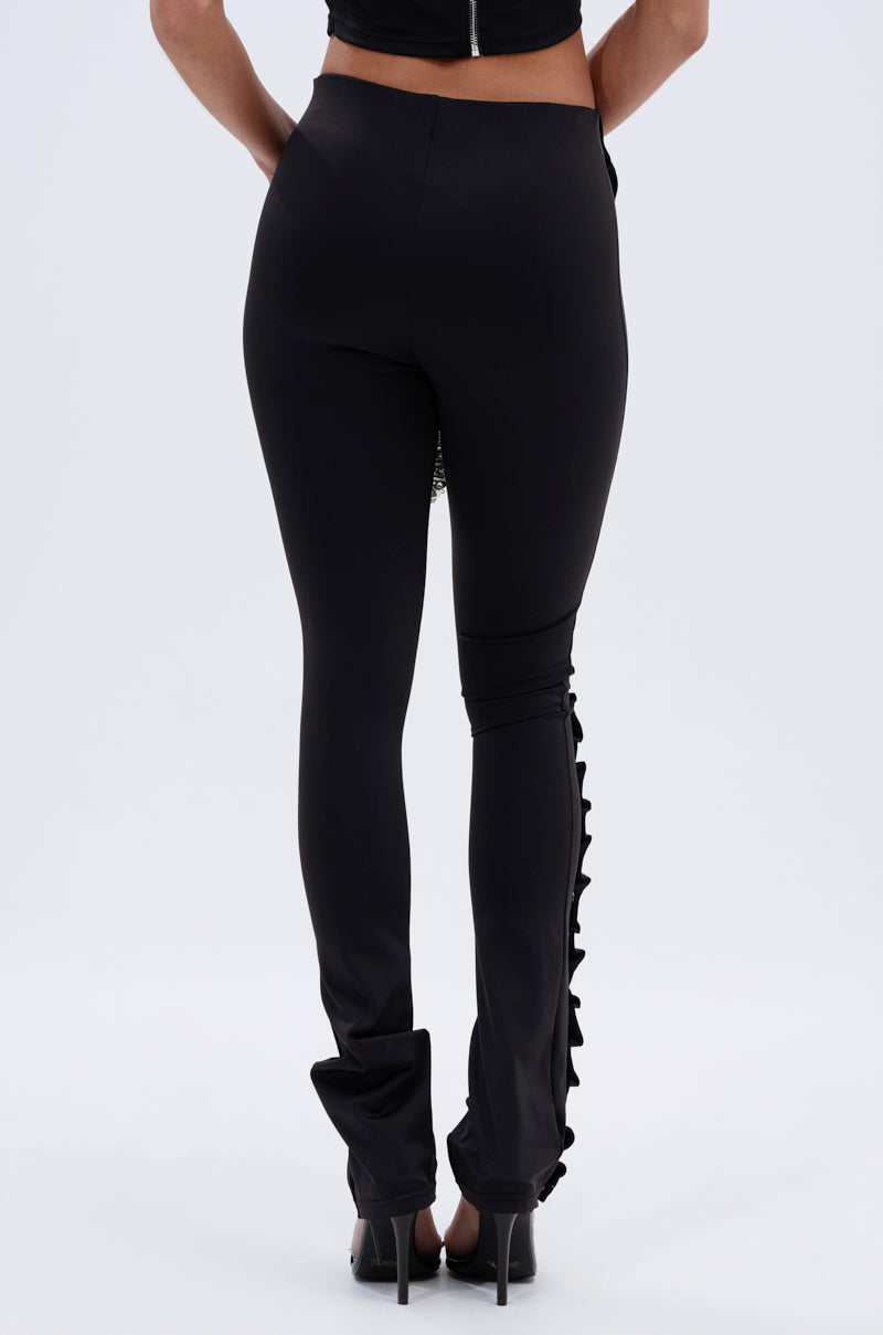 Someone wears the GROMMET DETAIL RUCHED PANT, featuring high-rise skinny fit with ruffle detail along one leg, paired with black high-heeled shoes, shown from the back against a light gray background. 4