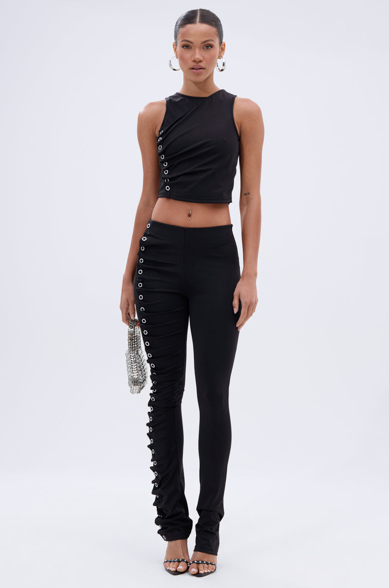 A woman faces forward in the GROMMET DETAIL RUCHED PANT—black, curve-hugging with side grommet details and leg ruching—paired with a black sleeveless crop top, heeled sandals, and a beaded handbag. 5