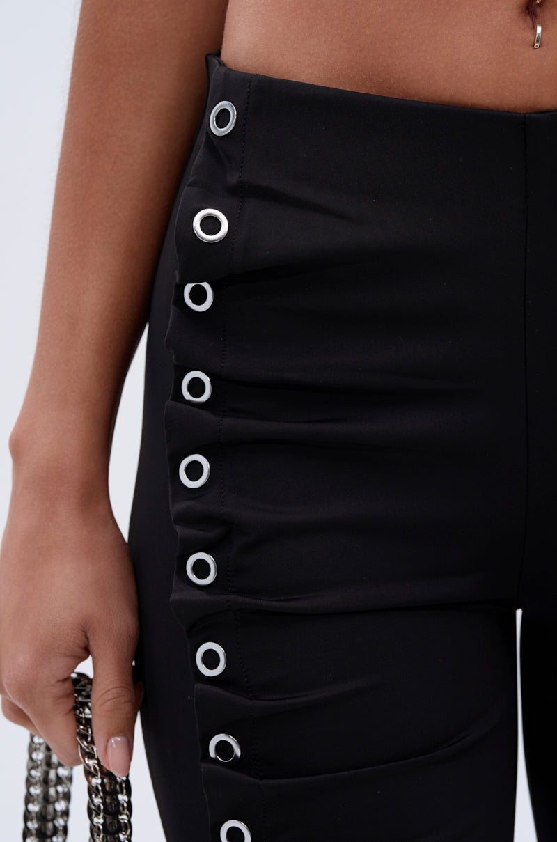 Close-up of a person wearing the GROMMET DETAIL RUCHED PANT, featuring high-rise, curve-hugging fabric and silver grommets along the side seam. The person holds a silver chain accessory in one hand. 6