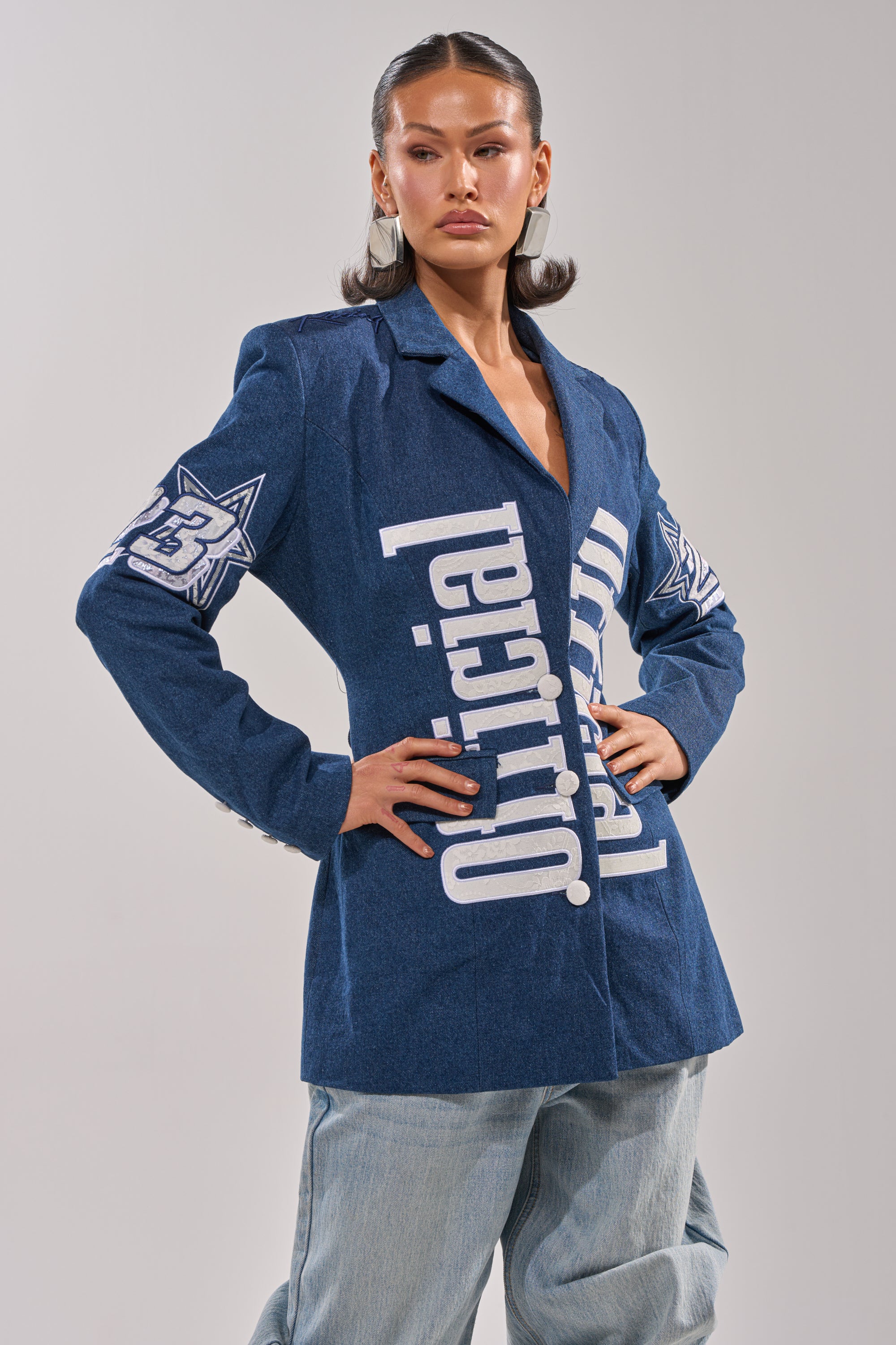 A woman with slicked-back hair and a serious expression models the ONE OF ONE BACKLESS DENIM BLAZER, featuring bold white text and star graphics, paired with light blue jeans against a plain gray background. 1
