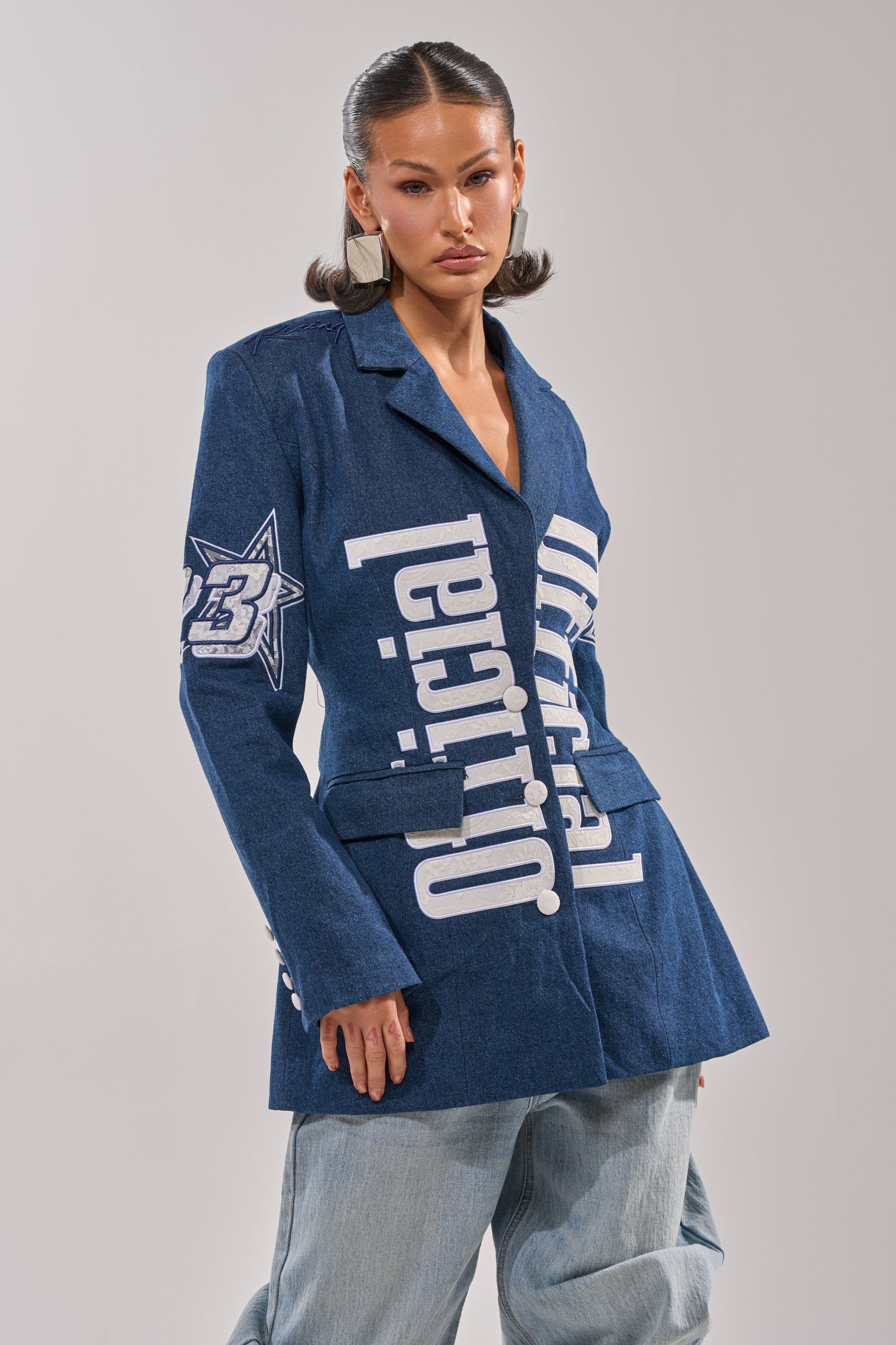 A woman models the ONE OF ONE BACKLESS DENIM BLAZER, featuring bold white text and graphic accents, paired with loose light blue jeans. Her brown hair is styled and she wears statement earrings, posing confidently against a plain background. 0