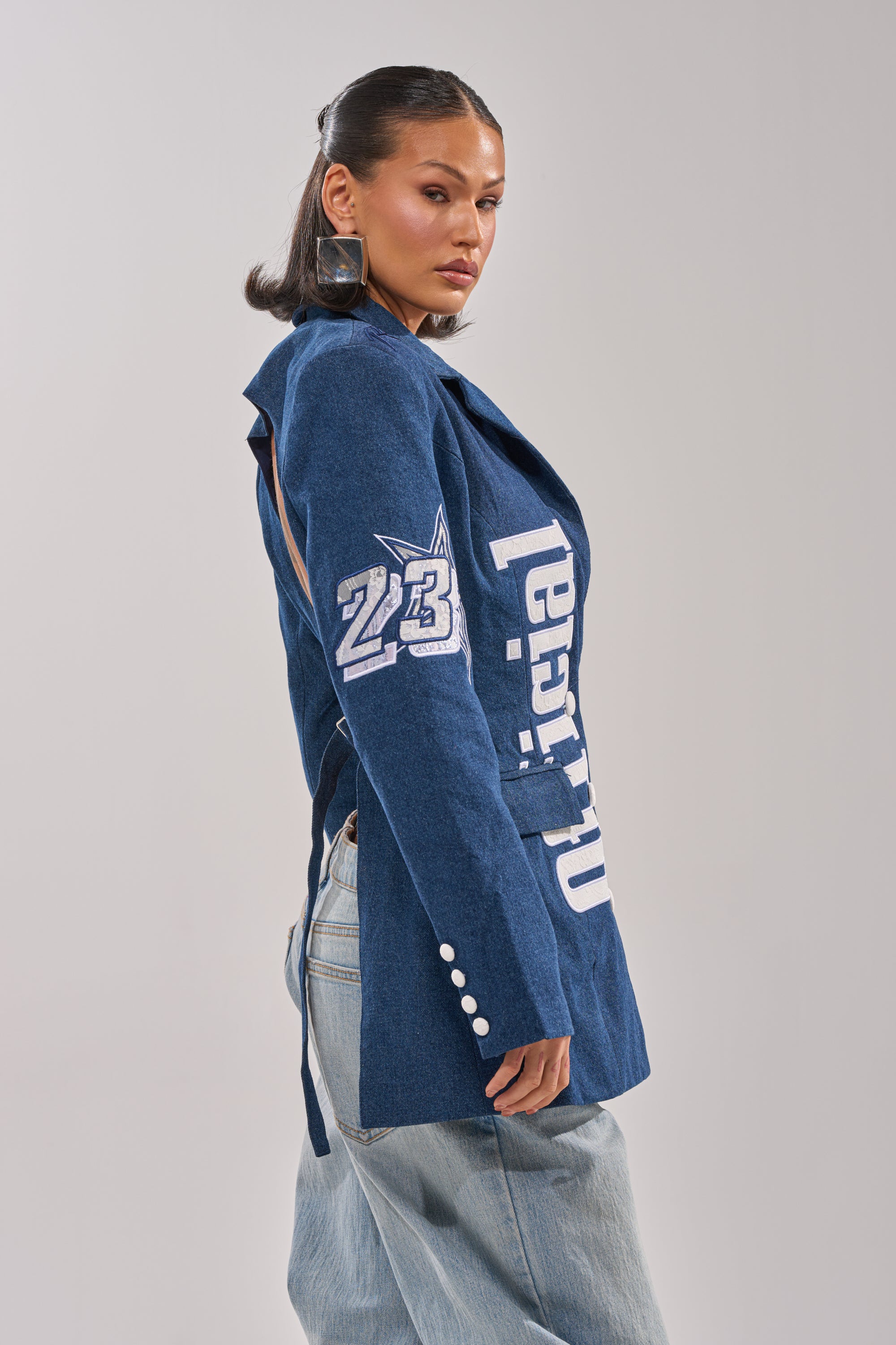 A woman poses in profile wearing the ONE OF ONE BACKLESS DENIM BLAZER, featuring white lettering and the number 23 on the sleeve, styled with light blue jeans and bold square earrings against a plain background. 2