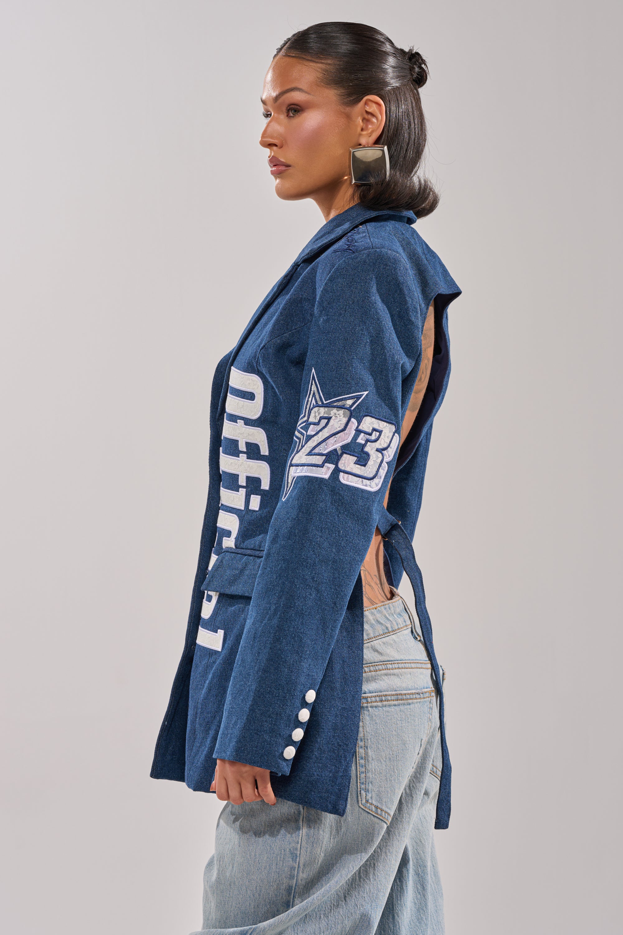 A woman in the ONE OF ONE BACKLESS DENIM BLAZER, featuring "Official" and "23" patches, stands in profile. Styled with light blue jeans and bold square earrings, she creates a standout streetwear look. 3
