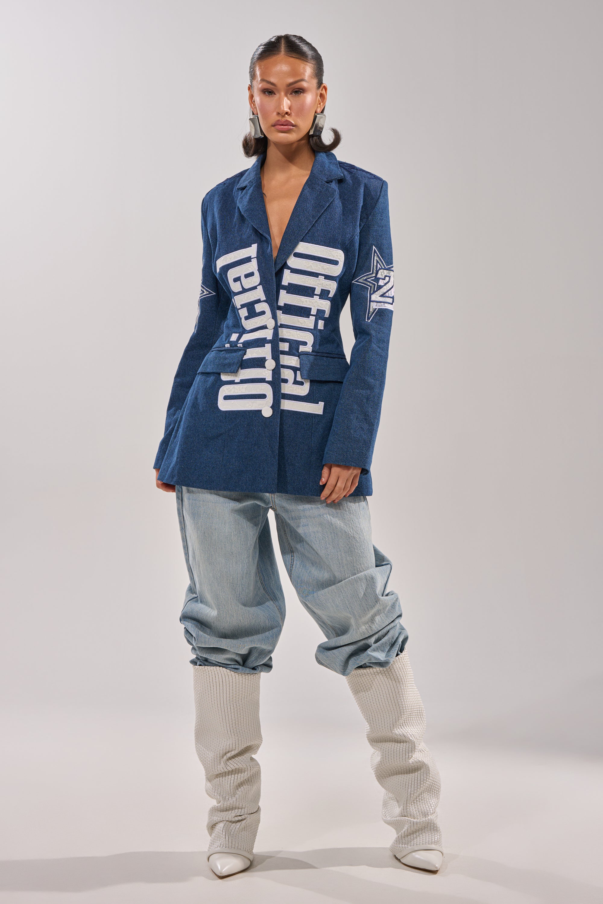 A woman exudes statement streetwear vibes in the ONE OF ONE BACKLESS DENIM BLAZER, paired with baggy light blue jeans tucked into tall off-white boots, posing confidently against a plain, light-colored background. 6