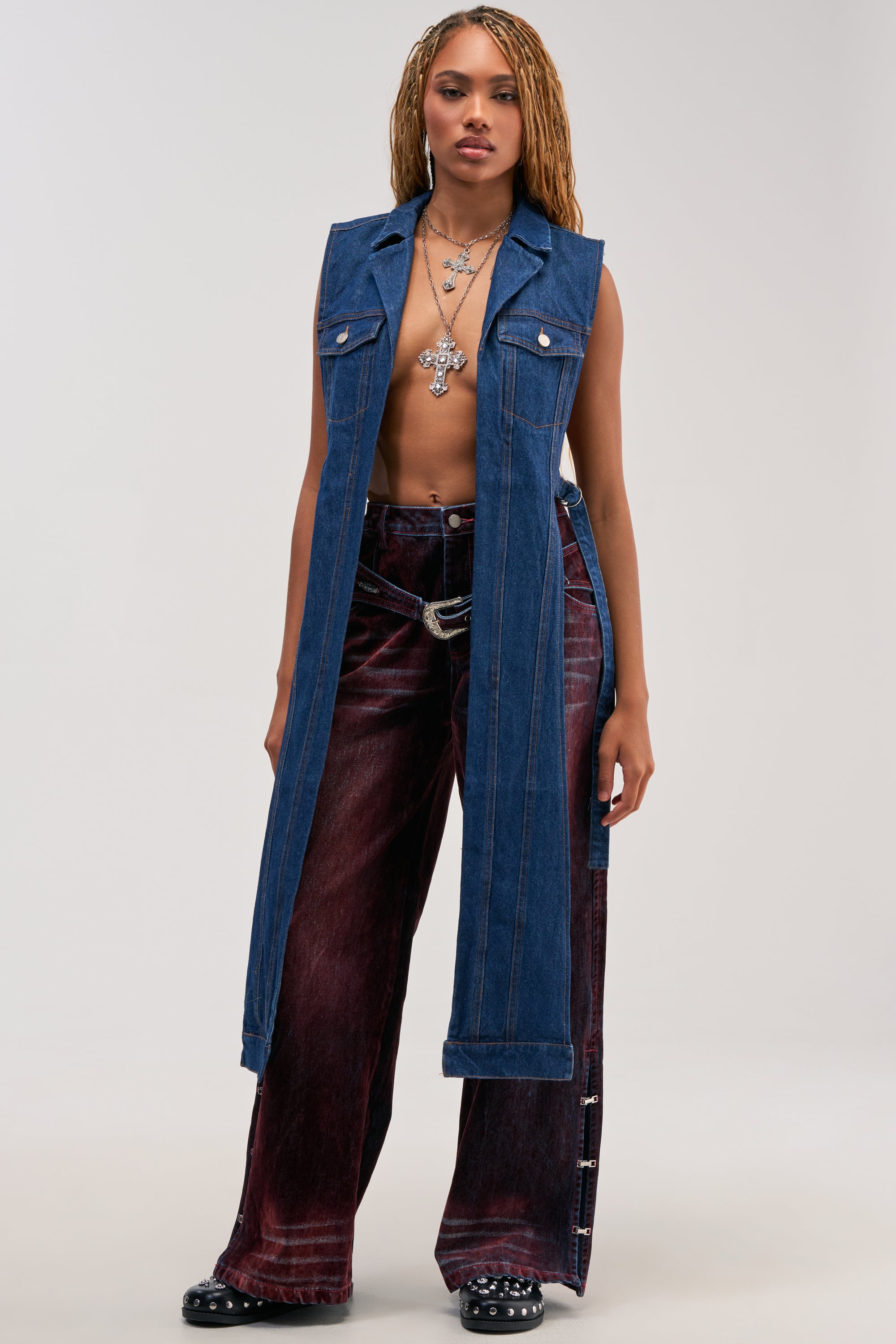 Someone with long, braided hair models the DOWN TO IT SPLIT VEST IN DARK DENIM—a tailored sleeveless denim vest—paired with maroon wide-leg jeans, black shoes, and layered necklaces. They pose confidently against a plain, light background.