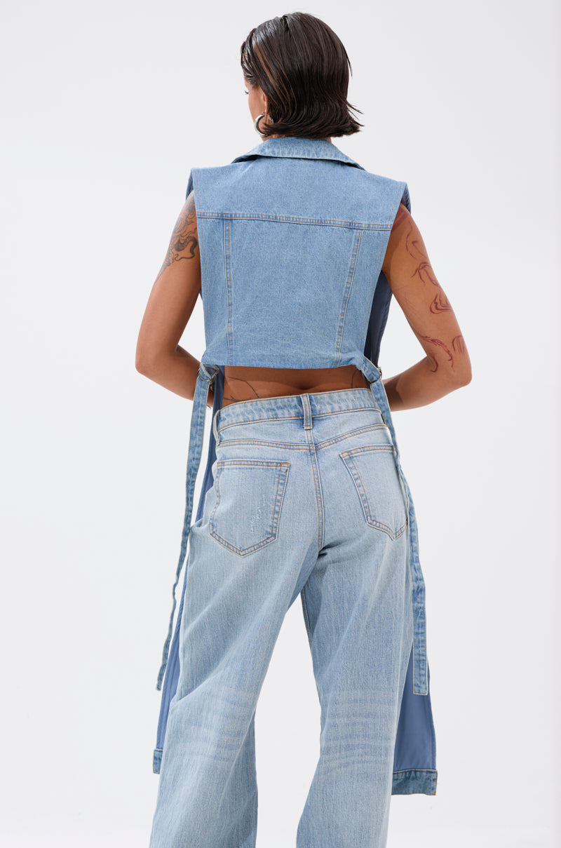 Model with short dark hair and visible tattoos faces away, wearing the DOWN TO IT SPLIT DENIM VEST styled over light blue jeans, showcasing modern denim layering against a plain white backdrop. 3