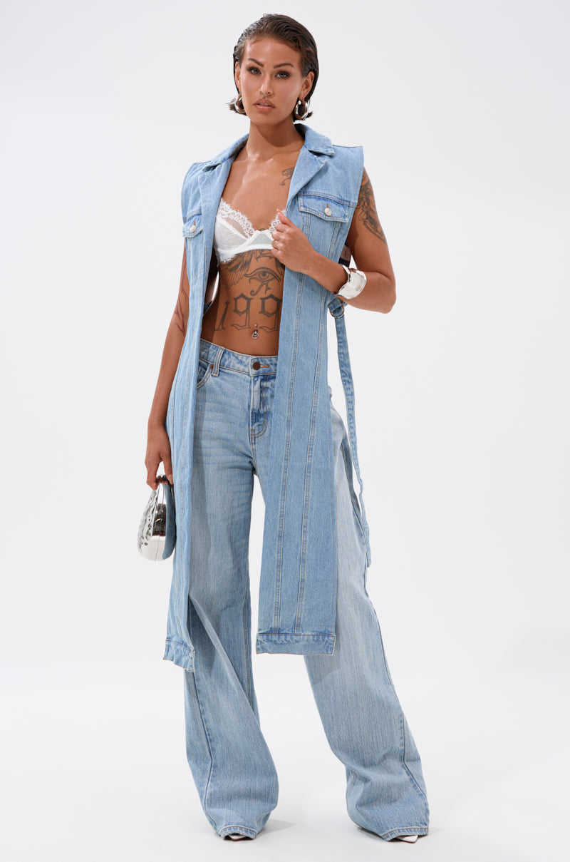 A person styles the DOWN TO IT SPLIT DENIM VEST over a white lace bralette and wide-leg jeans, paired with a white clutch, jewelry, and white shoes for a streetwear-inspired look against a plain white background. 4