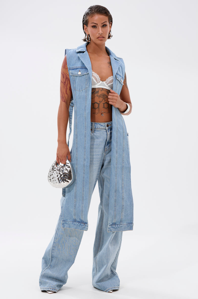 A woman stands confidently in the DOWN TO IT SPLIT DENIM VEST layered over a white lace bralette, paired with wide-leg jeans and white heels. She holds a small silver handbag, revealing tattoos on her left arm against a plain white background. 5