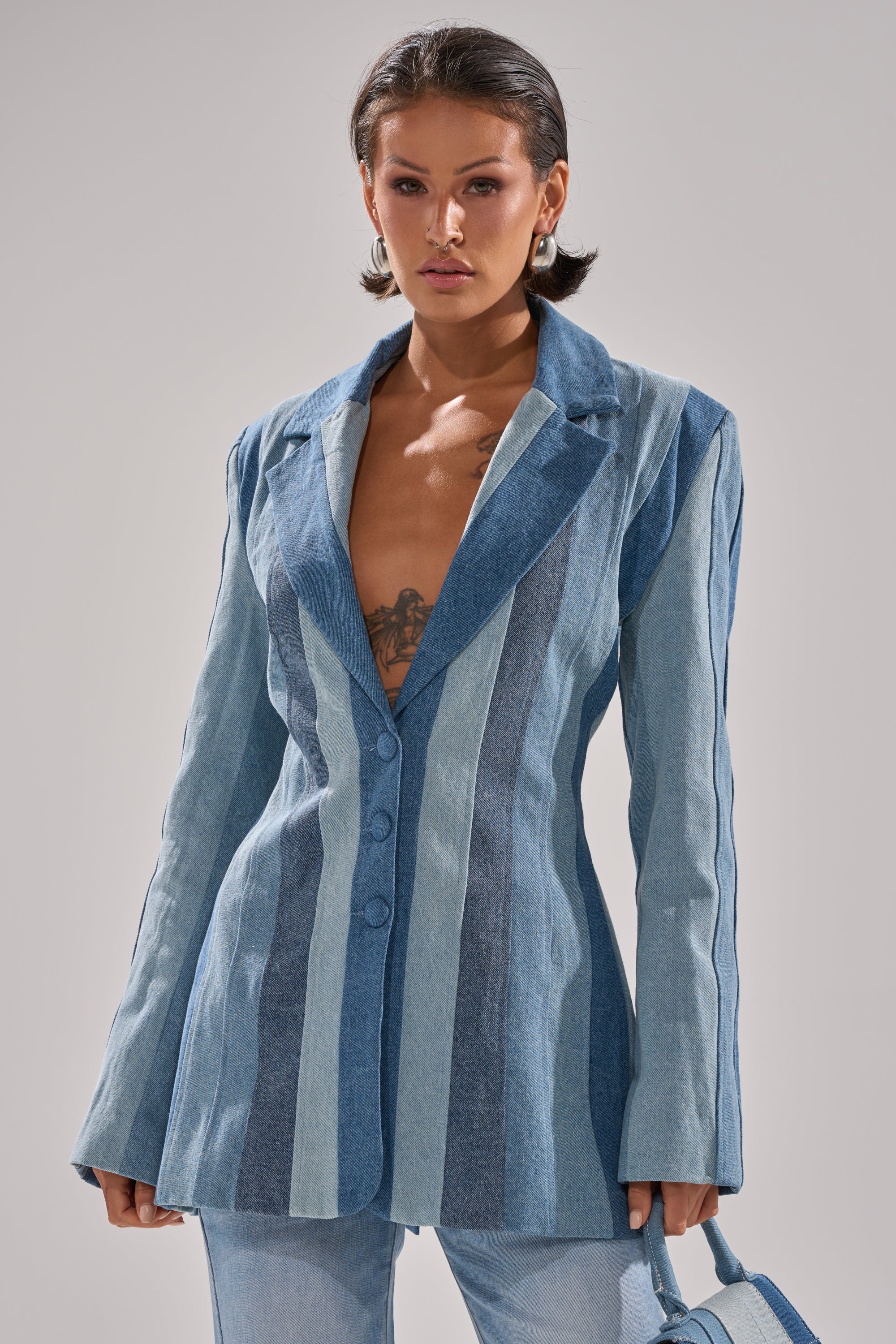 A woman with short dark hair wears the COME SLIDE PATCHWORK DENIM BLAZER without a shirt, revealing a chest tattoo. She pairs it with light blue jeans and holds a matching denim bag against a plain, light-colored background.