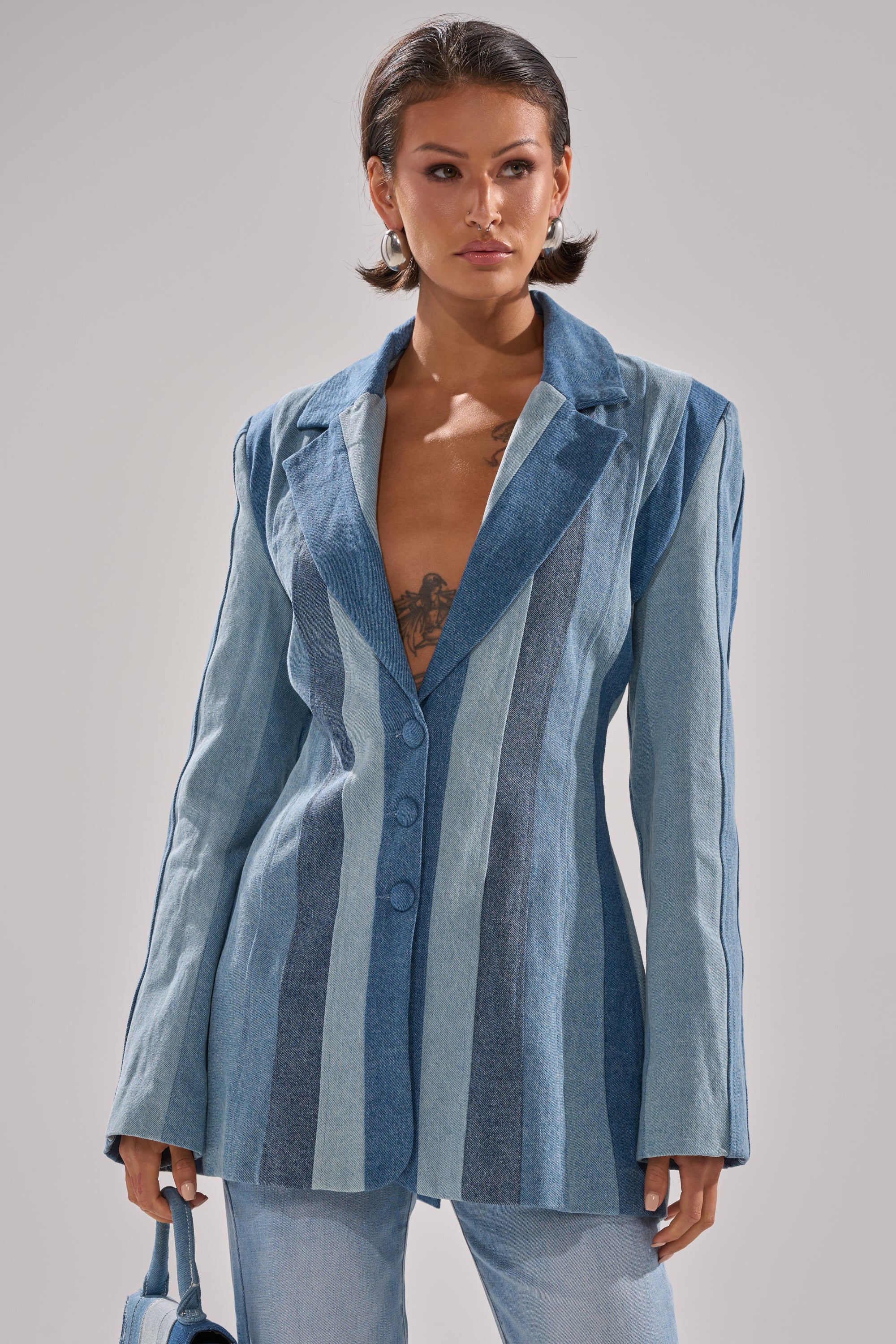 A woman in the COME SLIDE PATCHWORK DENIM BLAZER pairs it with light blue jeans and a small blue bag. Her short dark hair and visible chest tattoo add edge to her chic style.