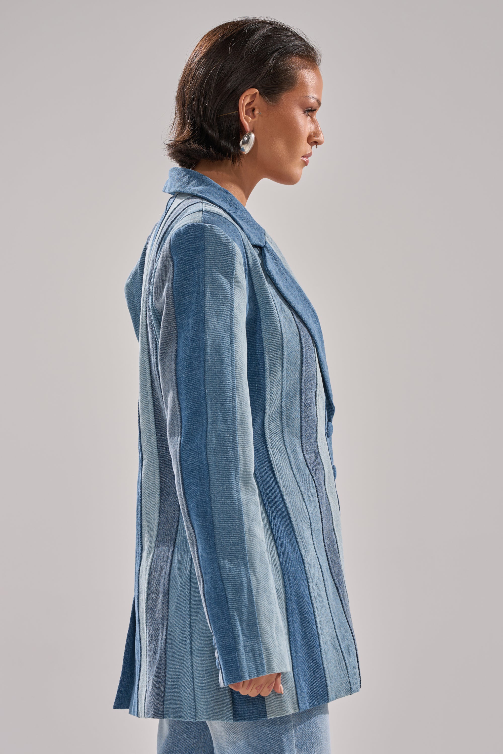 A person with short dark hair and silver hoop earrings models the COME SLIDE PATCHWORK DENIM BLAZER, standing in profile against a plain light gray background.