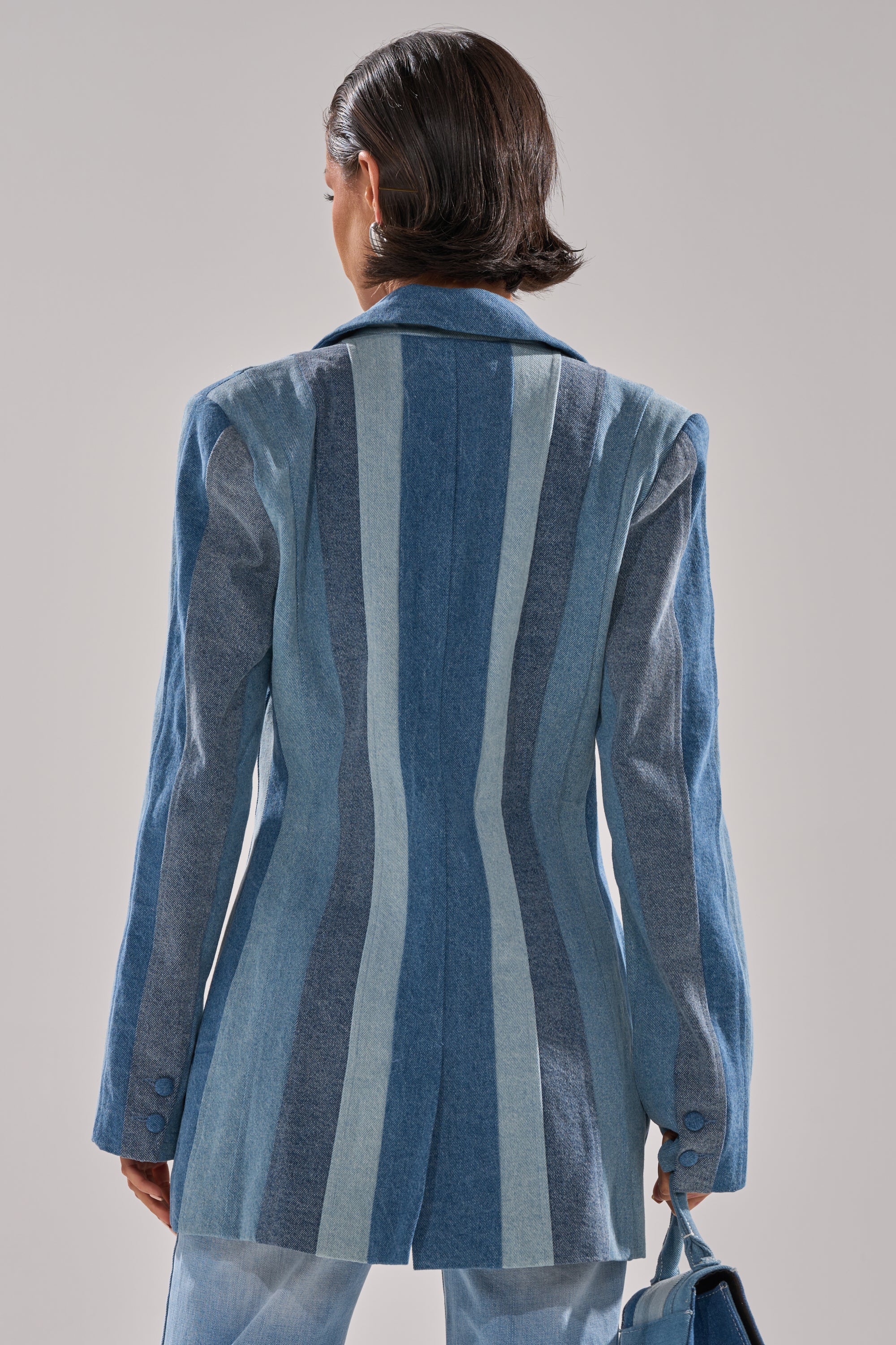 A person with short dark hair, seen from the back, wears the COME SLIDE PATCHWORK DENIM BLAZER with vertical blue and grey stripes, pairs it with light blue pants, and carries a matching blue bag.