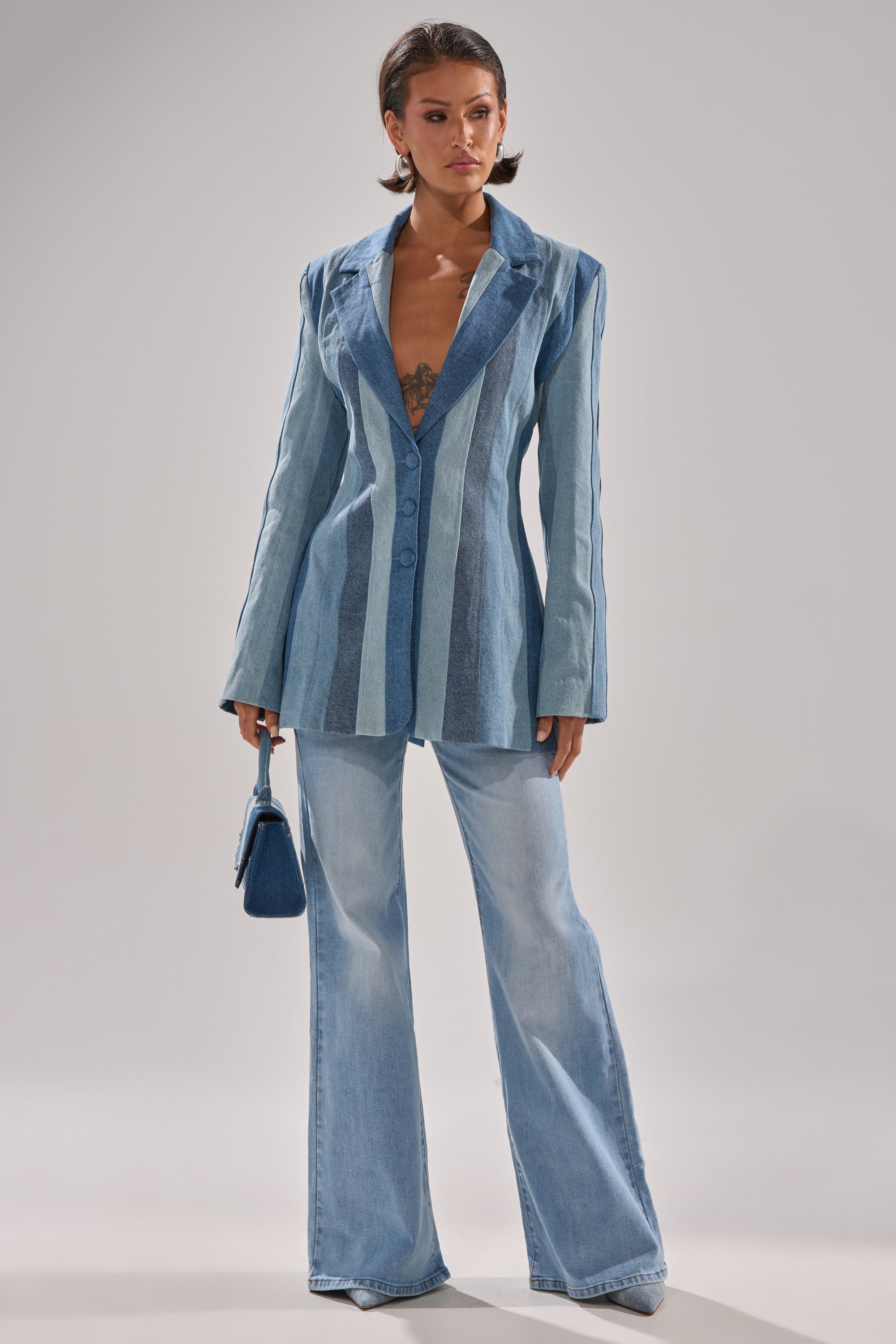 A woman stands confidently in the COME SLIDE PATCHWORK DENIM BLAZER, paired with flared light blue jeans and a small blue handbag. She sports short, styled hair and displays visible chest tattoos.