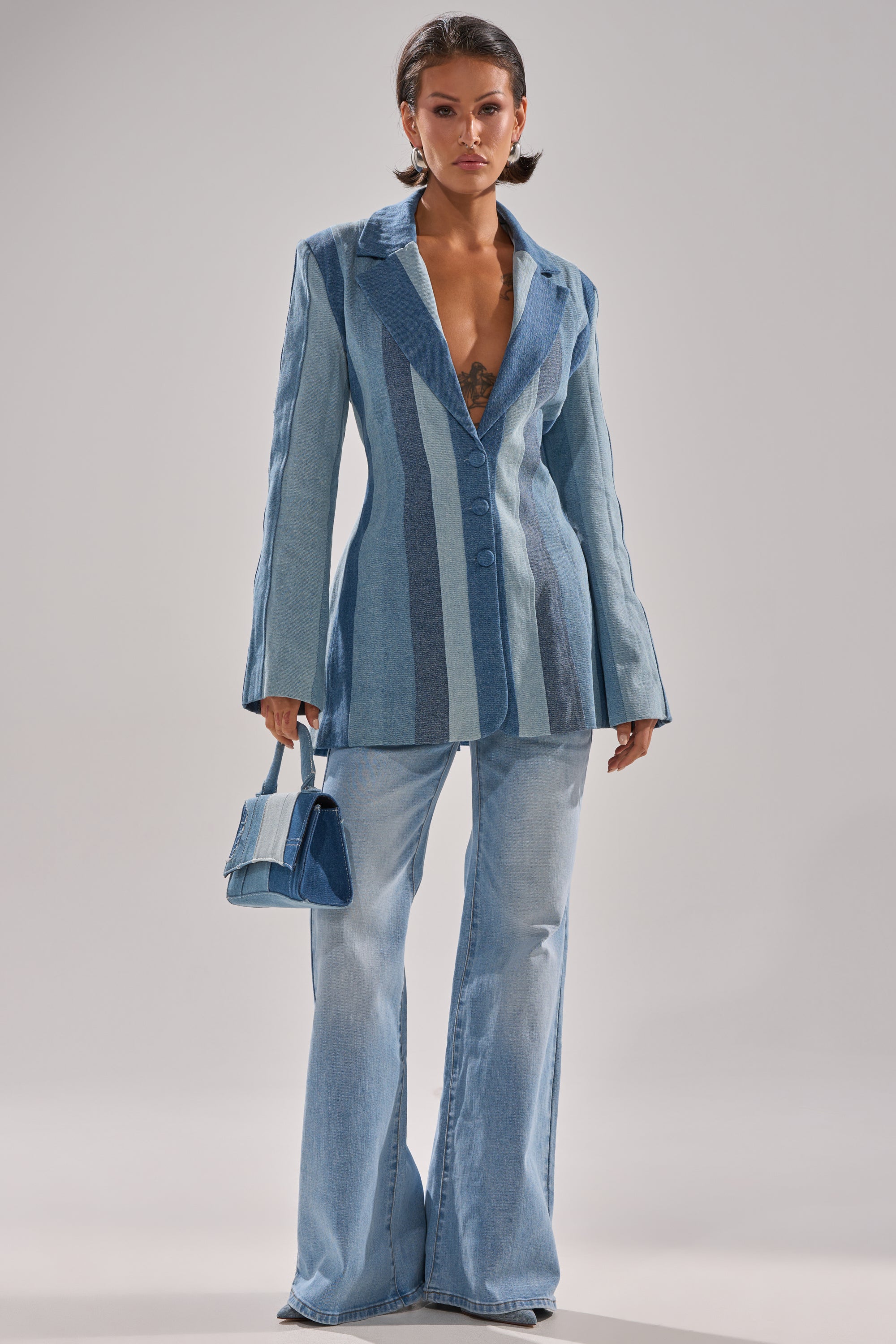 A woman with short dark hair confidently poses in the COME SLIDE PATCHWORK DENIM BLAZER, styled with light flared jeans and a small matching blue handbag.