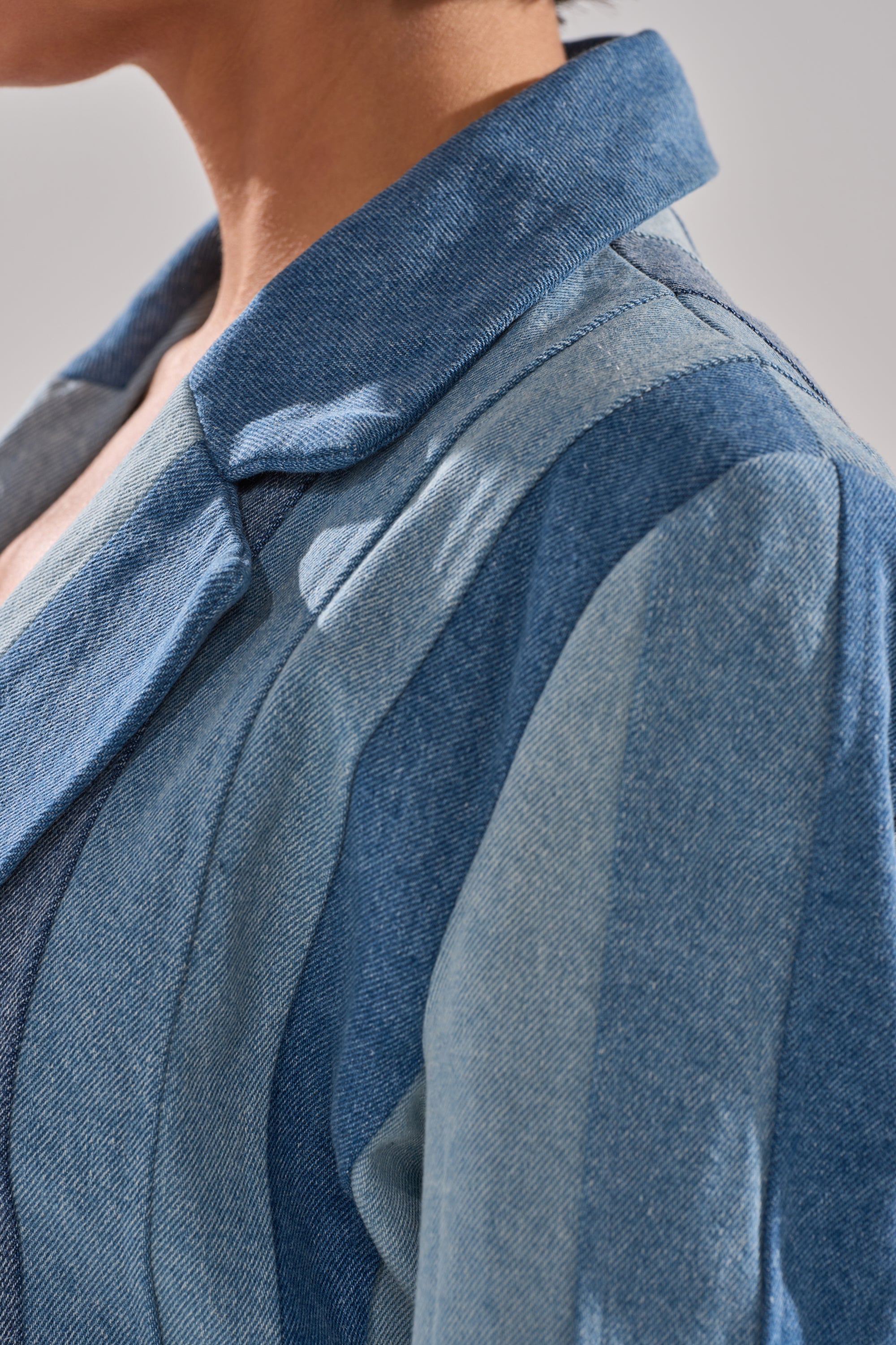 Close-up of a person wearing the COME SLIDE PATCHWORK DENIM BLAZER, featuring panels in varying shades of blue with detailed stitching and textured fabric. The person’s face is partly visible, turned to the side.