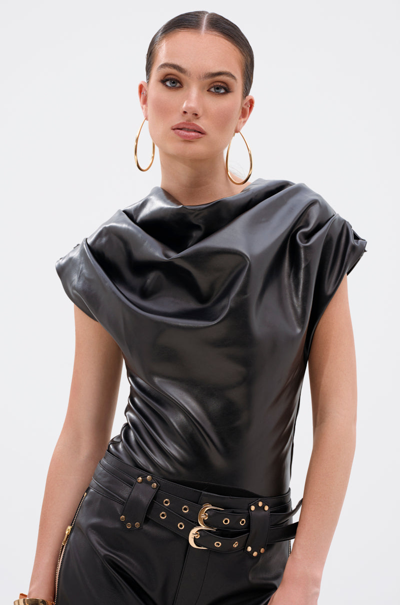 A woman poses against a plain white background wearing the SHANNON FAUX LEATHER BODYSUIT, featuring shiny black vegan leather and a draped cowl neckline, styled with matching belted pants and large gold hoop earrings.