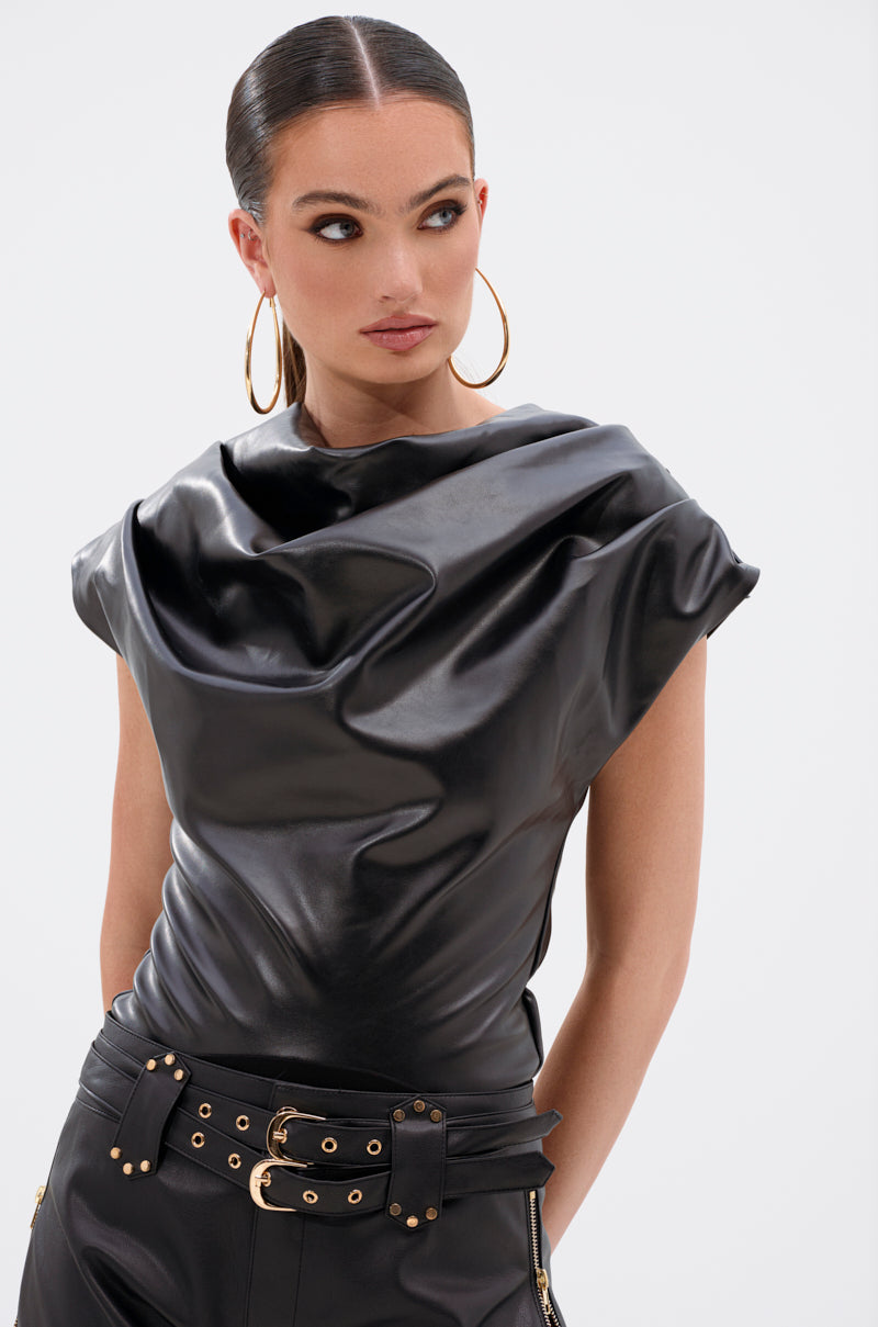 A woman in the SHANNON FAUX LEATHER BODYSUIT—sleek, black, draped, with a cowl neckline—pairs it with high-waisted pants and gold hardware. She wears hoop earrings and has slicked-back hair, standing against a plain white background looking to the side.