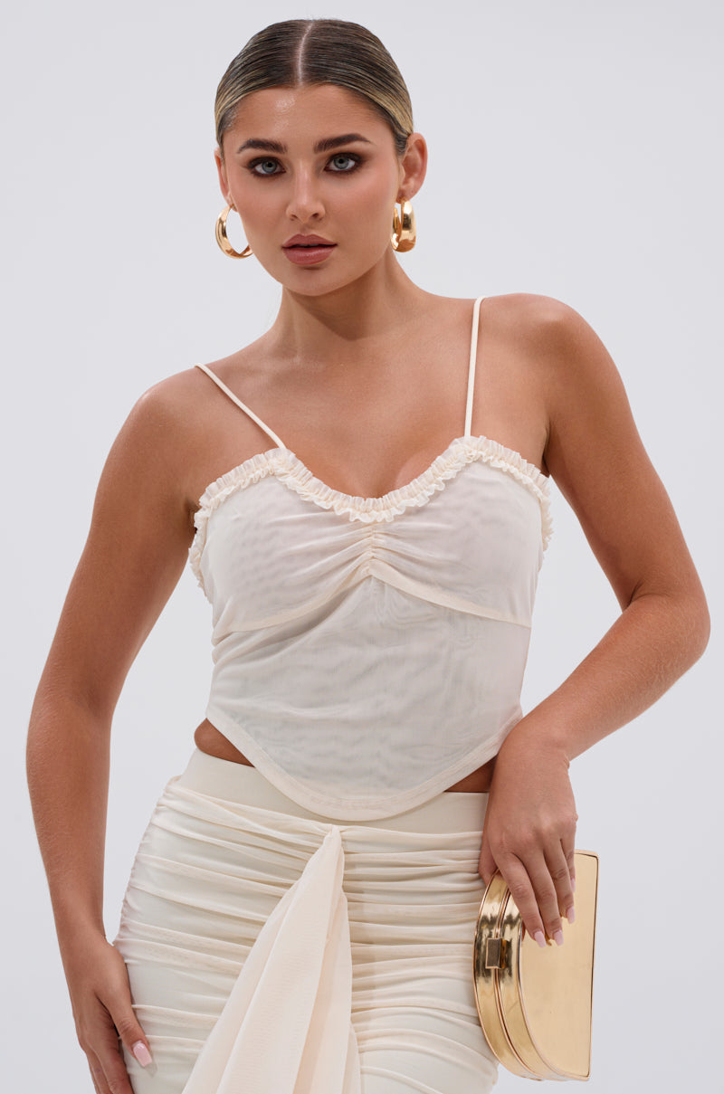 A woman with slicked-back hair wears the MELISSA MESH CAMI—a cream ruched bust spaghetti strap top with ruffle trim—paired with a matching skirt, gold hoop earrings, and a gold clutch, posed against a plain light background. 1