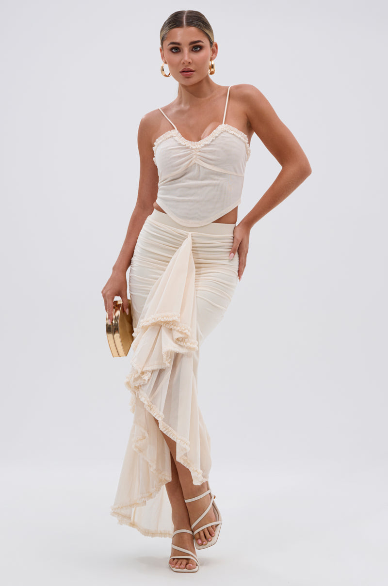 Wearing the MELISSA MESH CAMI and a cream ruffled maxi skirt, a woman holds a round clutch and poses confidently in strappy white heels against a plain white background. 4