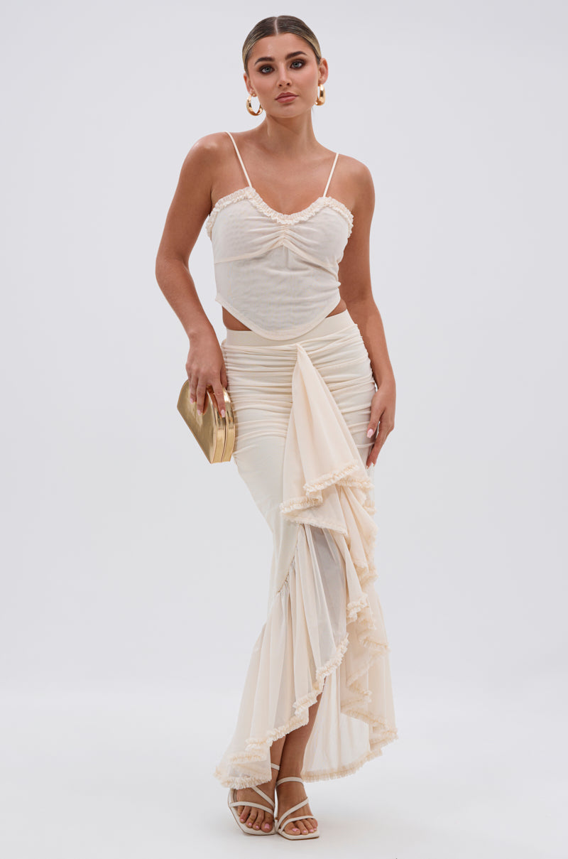A woman poses against a white background wearing the MELISSA MESH CAMI, a cream-toned ruched bust two-piece with spaghetti straps and a high-slit layered skirt. She accessorizes with gold hoop earrings and a matching clutch, hair sleekly styled back. 5