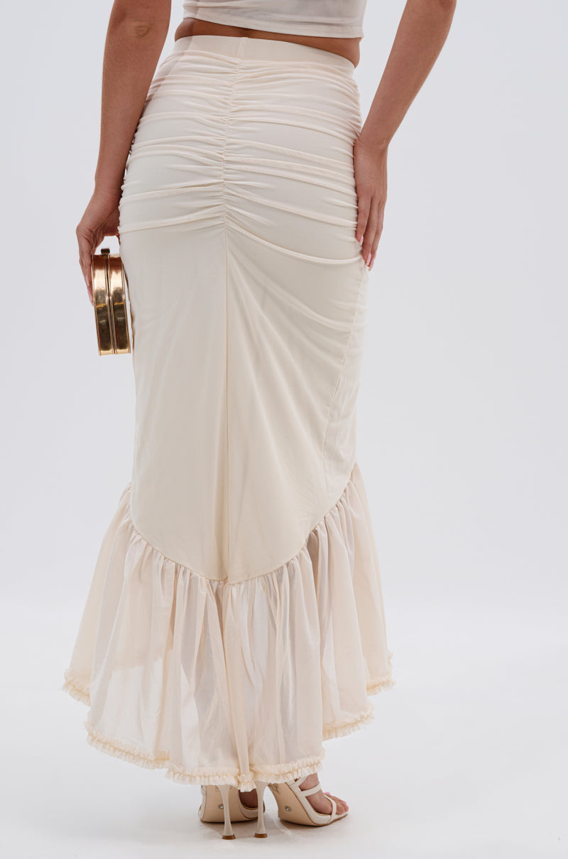 Wearing the MELISSA MESH RUCHED SKIRT with a fitted cream mesh design and sheer ruffled hem, this person pairs it with open-toe heels and a small round gold clutch while posing with their back to the camera. 3