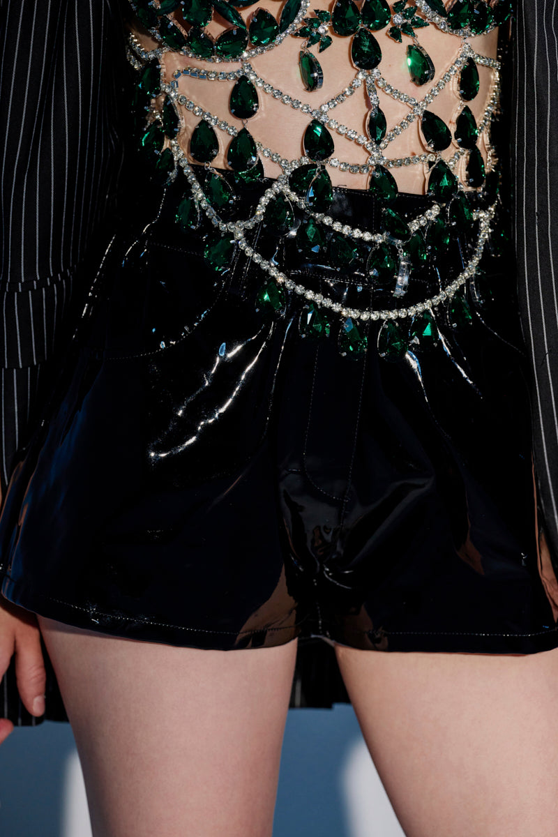 A close-up of a person wearing the DONT MESS WITH ME FAUX LEATHER SHORT paired with a top adorned with large green gemstones, silver chains, and pinstriped black fabric, revealing some skin. 0