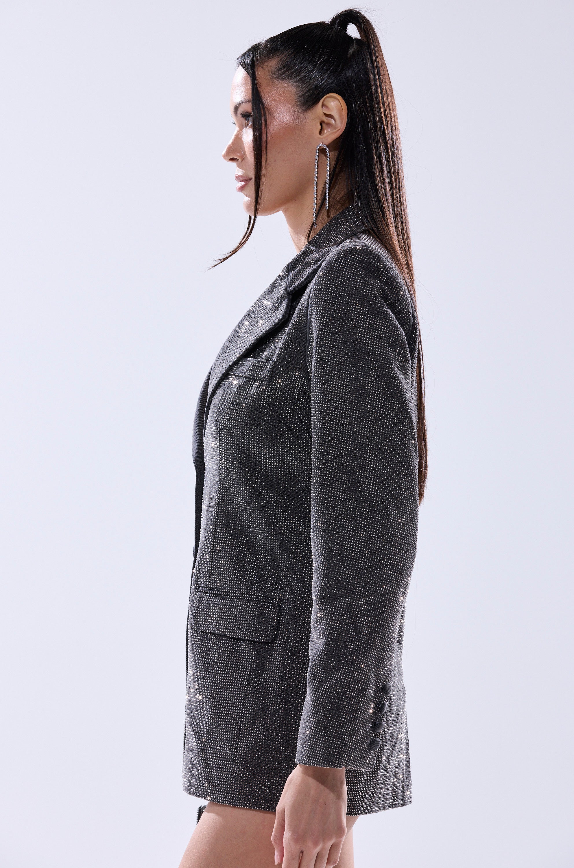 A woman with long, dark hair in a high ponytail models the ALL EYES ON ME EMBELLISHED DENIM BLAZER and long, dangling earrings as she stands in profile against a plain light background. 4